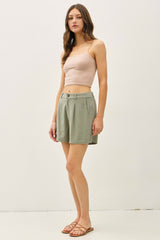 woman wearing a beige crop top and green high-waisted shorts, standing with a neutral background