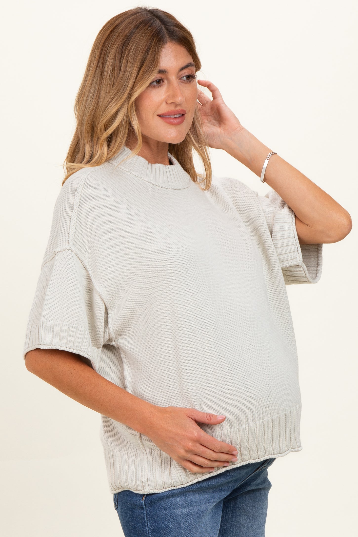 woman wearing a light gray oversized maternity sweater, gently cradling her baby bump with a relaxed expression