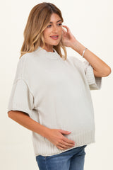 woman wearing a light gray oversized maternity sweater, gently cradling her baby bump with a relaxed expression