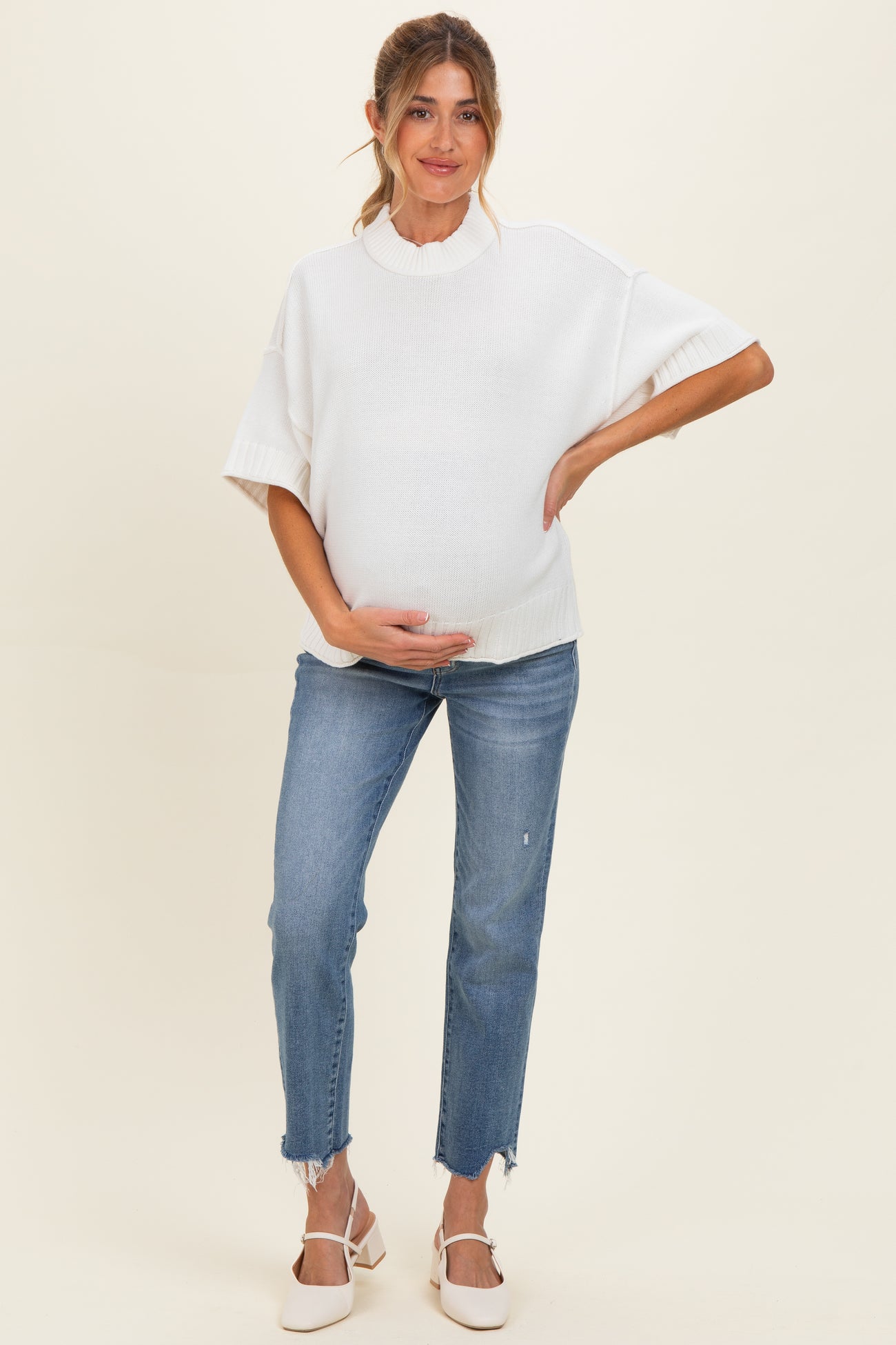 woman wearing a white short sleeve maternity sweater and blue distressed jeans, holding her baby bump