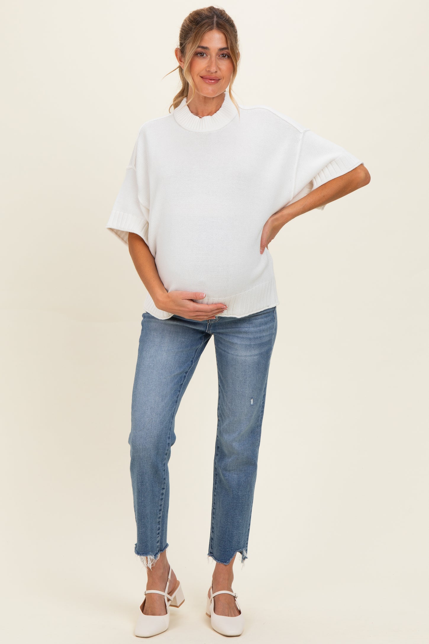 woman wearing a white short sleeve maternity sweater and blue distressed jeans, holding her baby bump