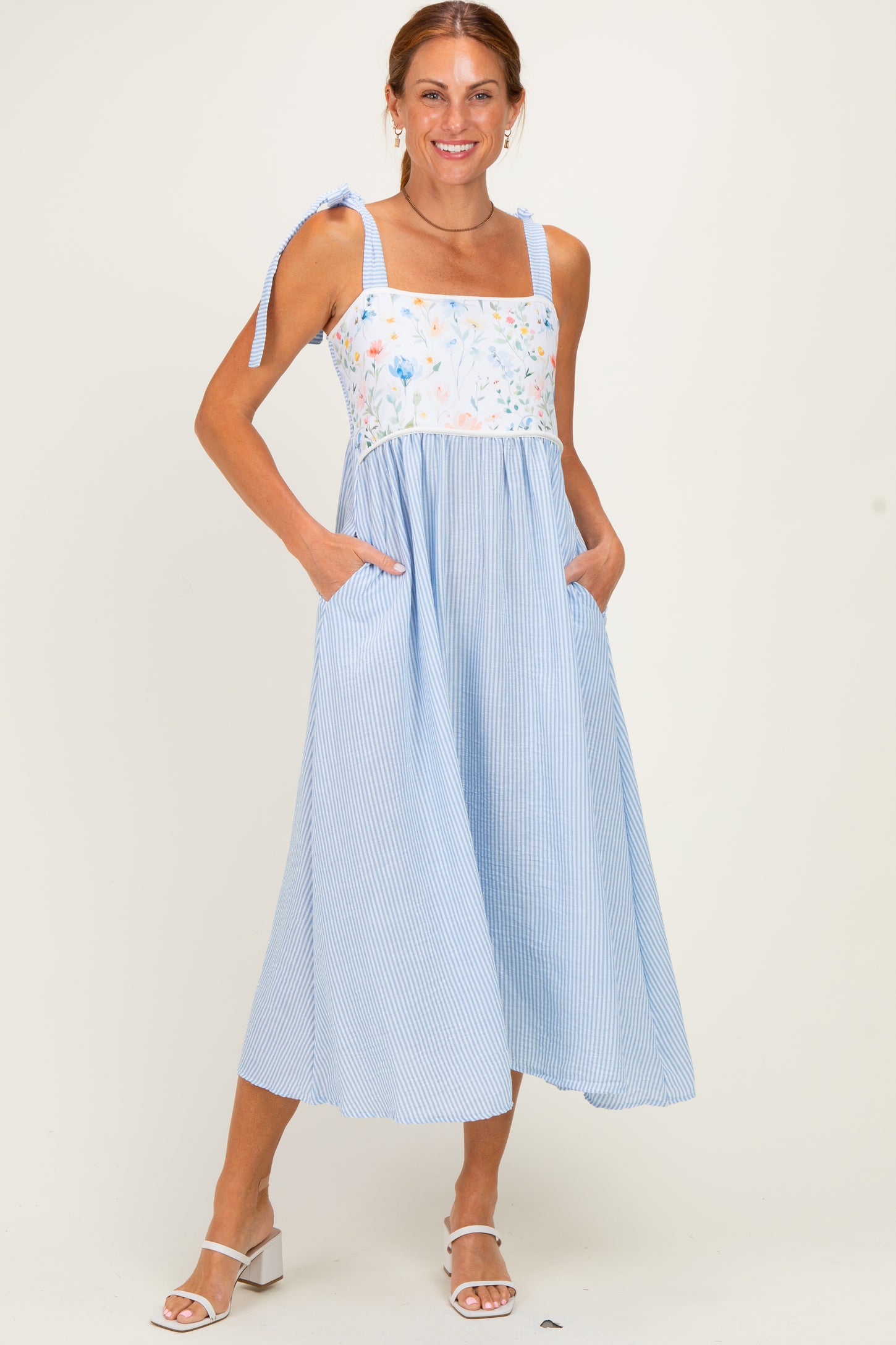 woman wearing a light blue striped sundress with floral bodice and pockets, standing on a neutral background
