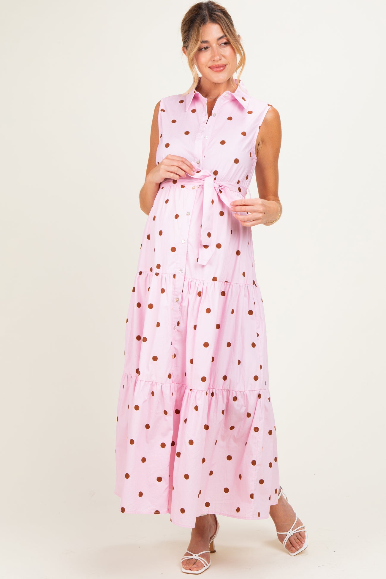 woman wearing a pink polka dot maxi maternity dress with a belted waist and sleeveless design, showing off her baby bump