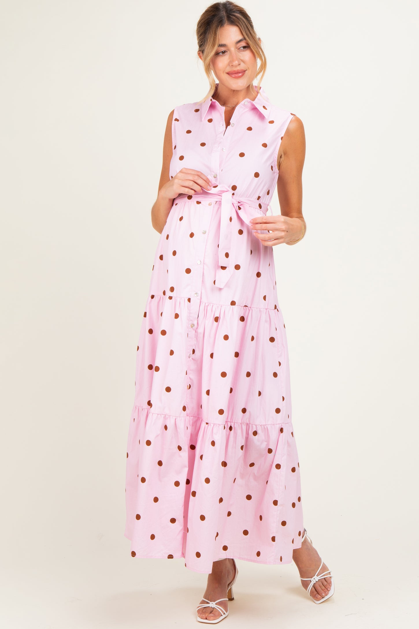 woman wearing a pink polka dot maxi maternity dress with a belted waist and sleeveless design, showing off her baby bump