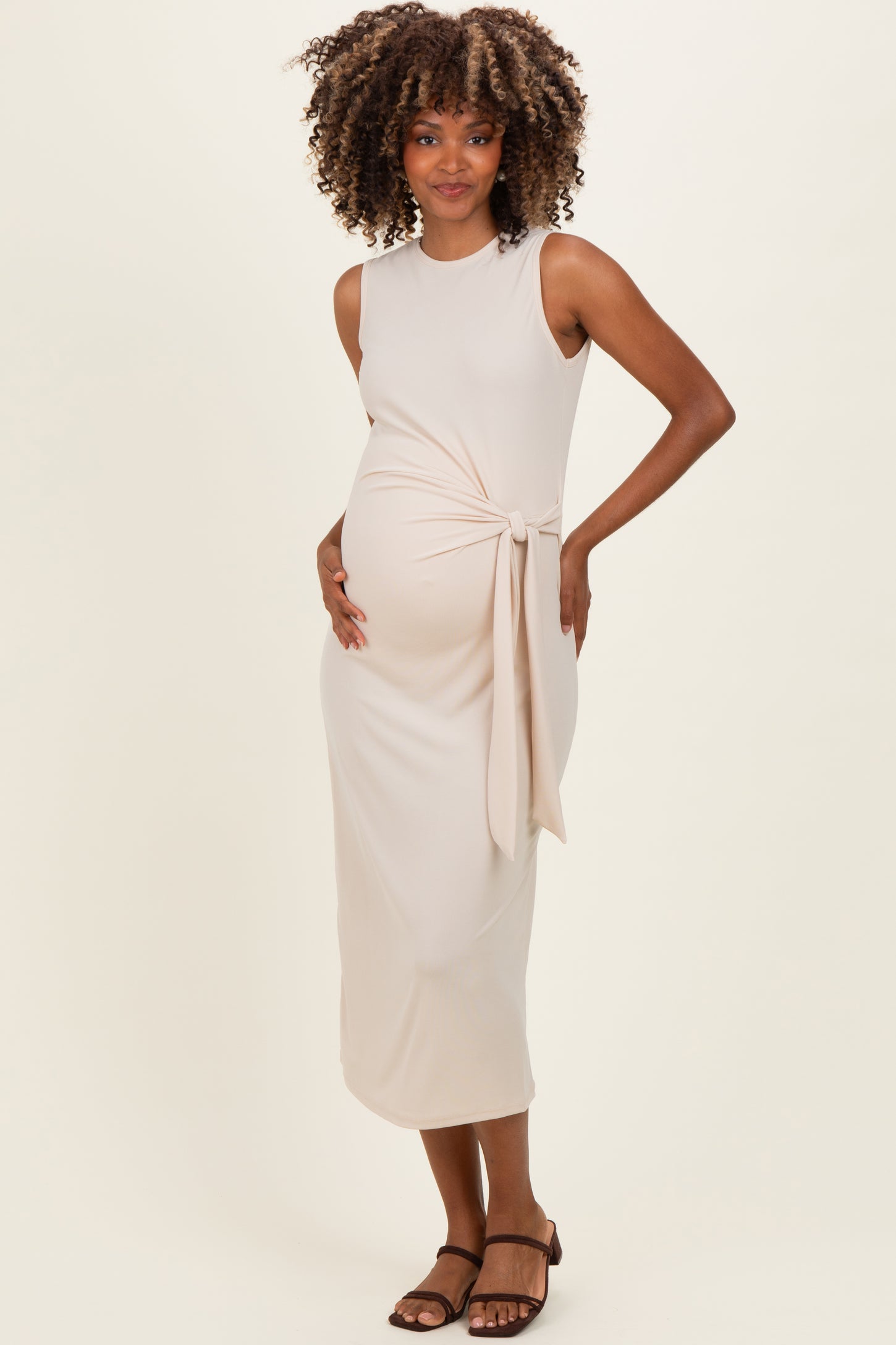 woman wearing a nude sleeveless maternity dress with a knotted detail and holding her baby bump