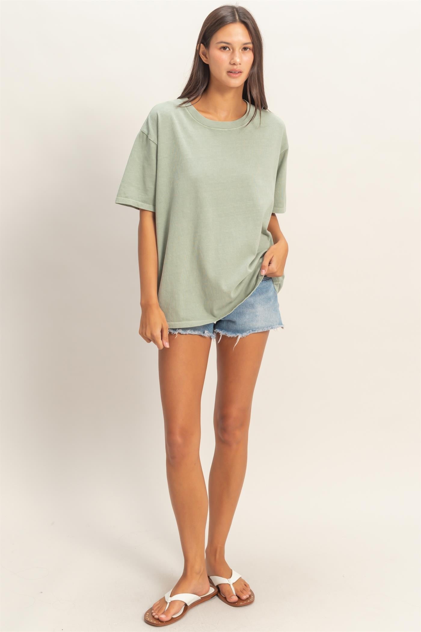 woman wearing an oversized sage green t-shirt and light blue denim shorts, standing barefoot in casual pose
