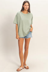 woman wearing an oversized sage green t-shirt and light blue denim shorts, standing barefoot in casual pose