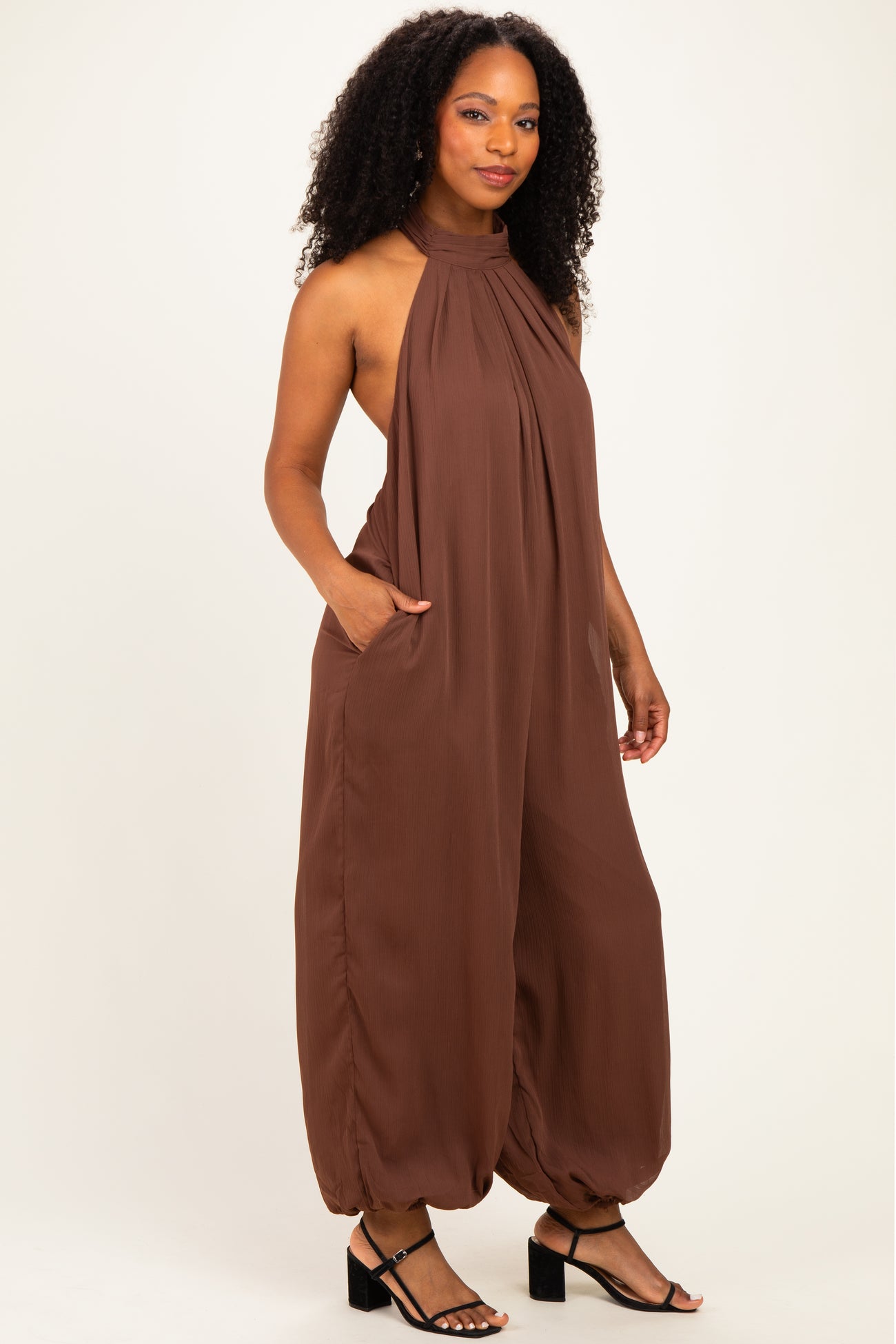 woman wearing a brown sleeveless jumpsuit with a halter neck and side pockets, styled with black strappy heels