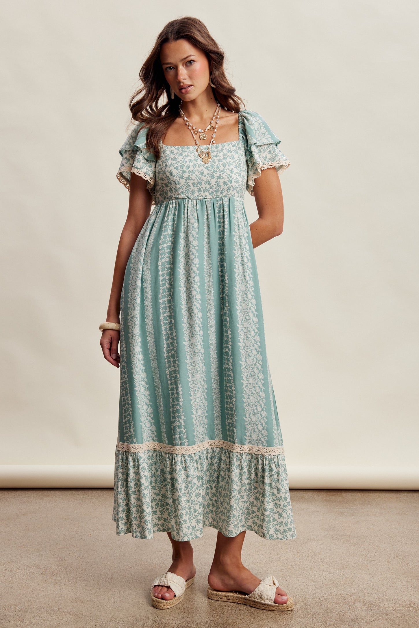 woman wearing a teal floral maxi dress with ruffled sleeves and lace trim, styled with sandals and layered jewelry
