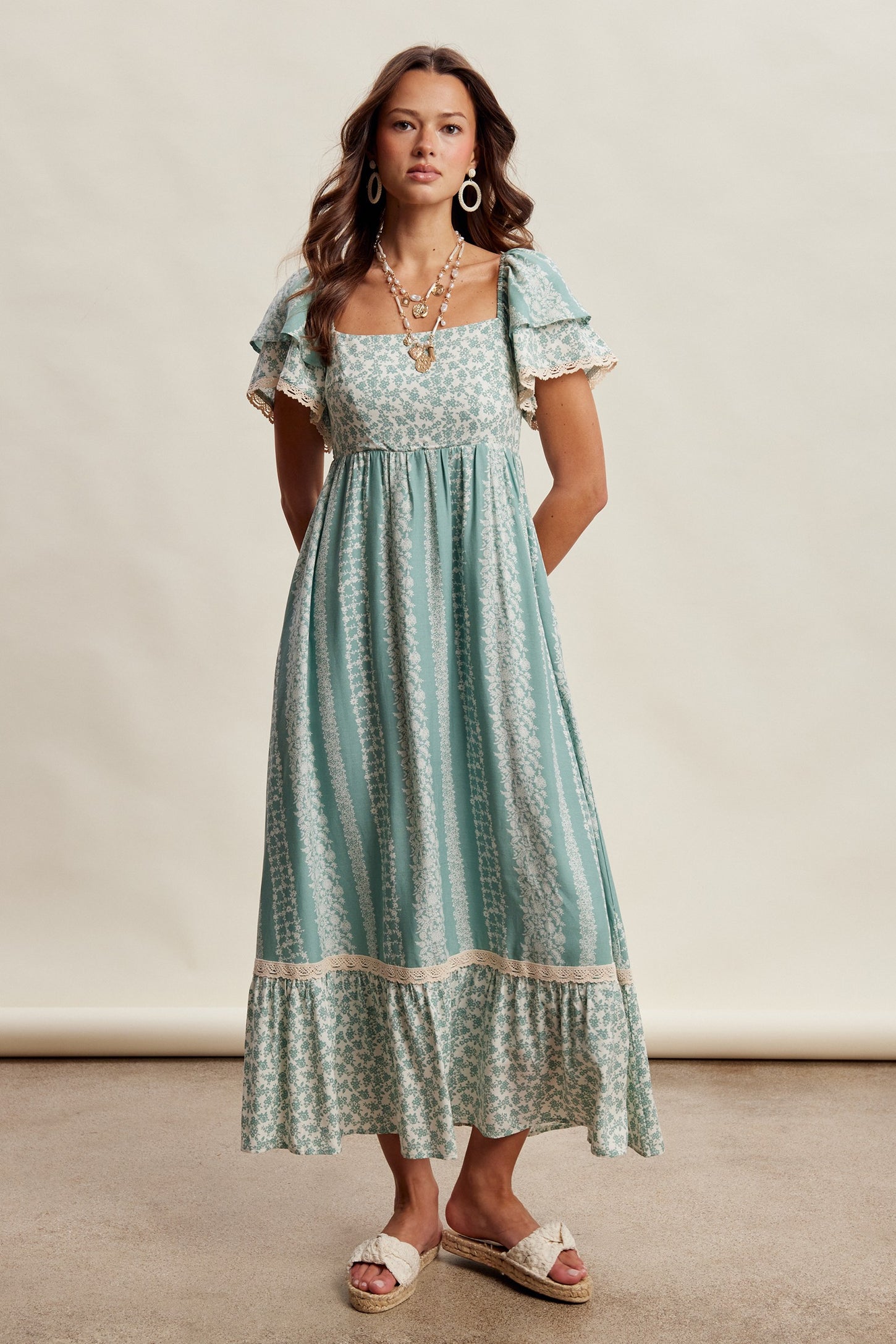woman wearing a teal floral maxi dress with short ruffled sleeves and lace trim, styled with layered necklaces and sandals