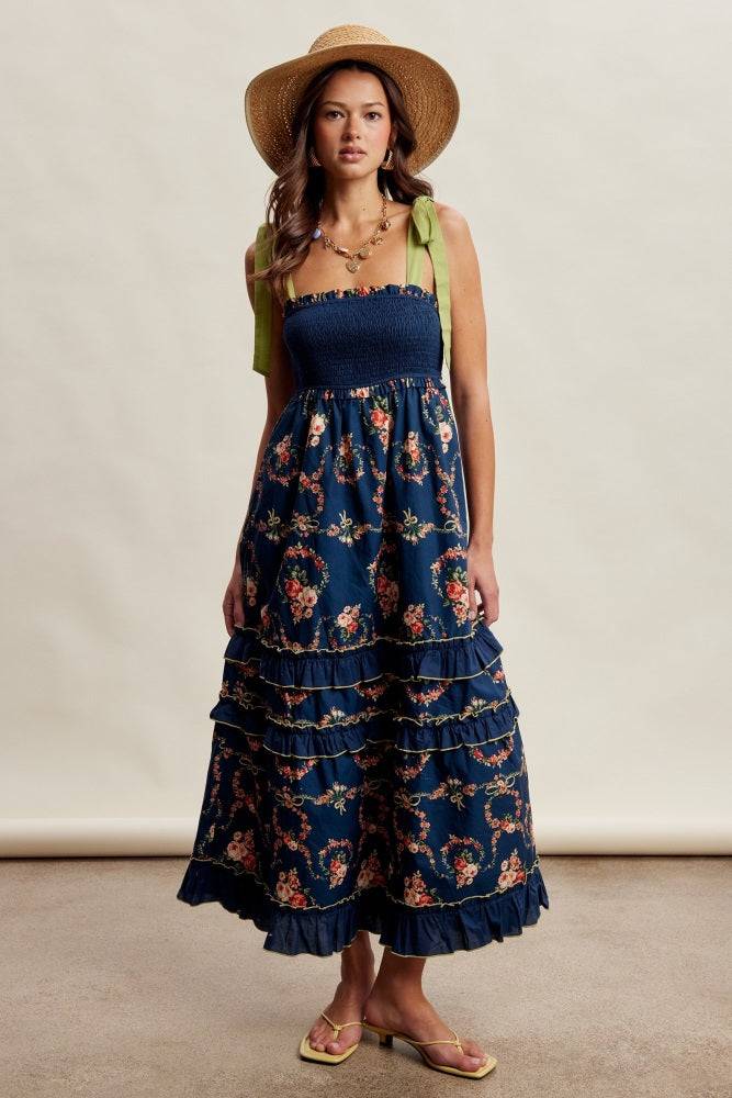woman wearing a navy blue floral maxi dress with ruffled tiers and green tie straps, accessorized with a straw hat
