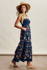 woman wearing a navy floral maxi dress with tiered ruffles and green shoulder straps, accessorized with a straw hat