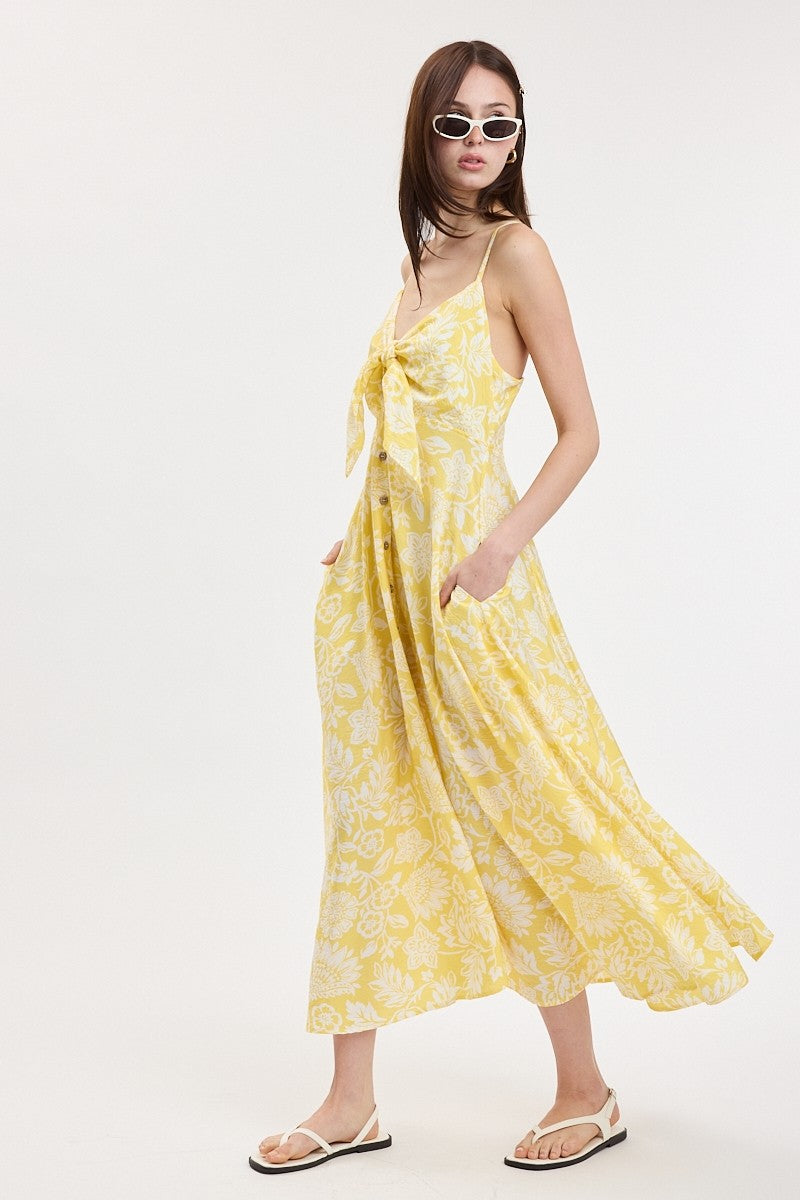 woman wearing a yellow floral print maxi dress with a tie front and side pockets, styled with white sunglasses and sandals