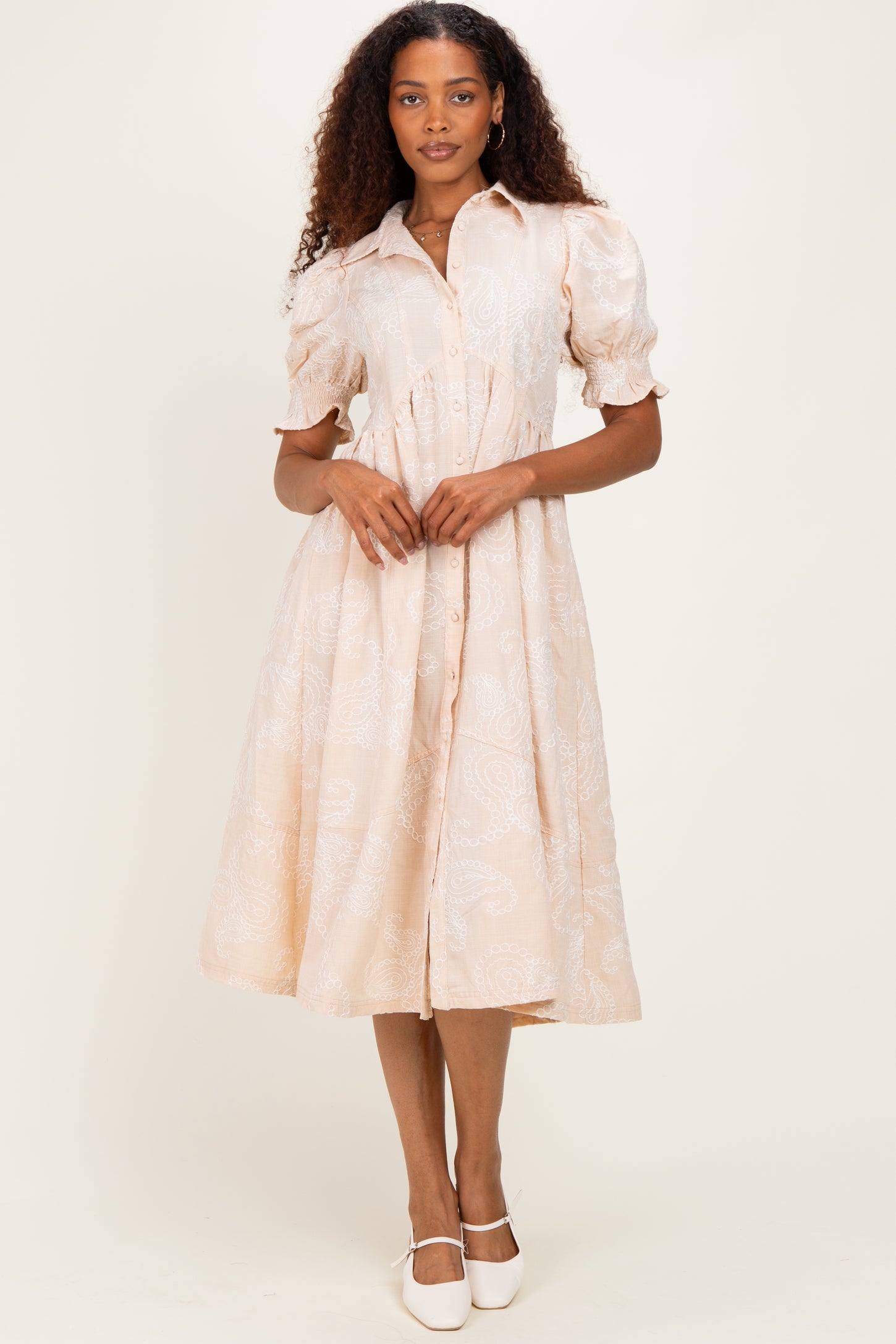 woman wearing a light peach button-up dress with puff sleeves and a paisley pattern, standing in a neutral background