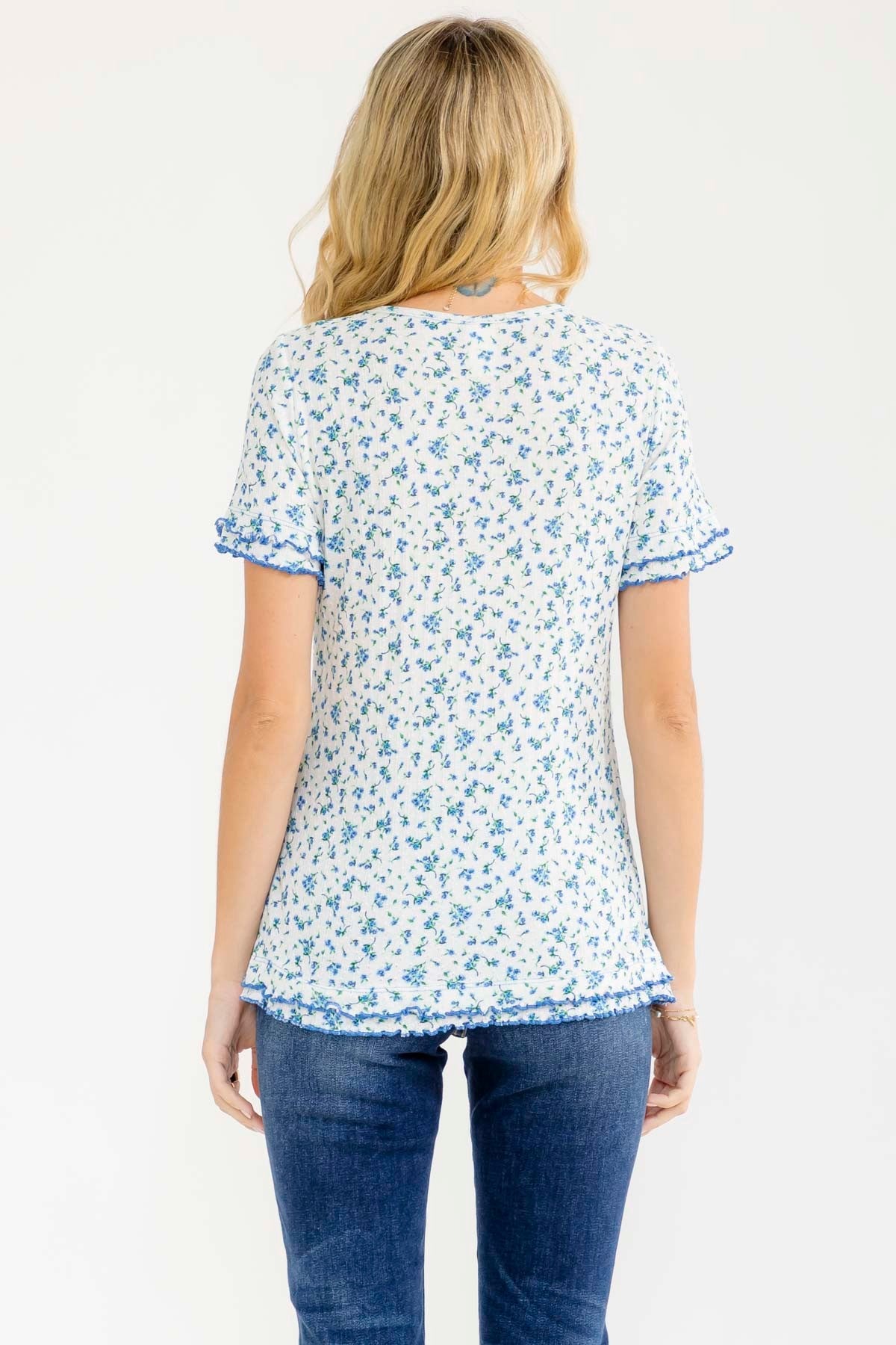 woman wearing a white short sleeve top with blue floral print and ruffle details, view from the back