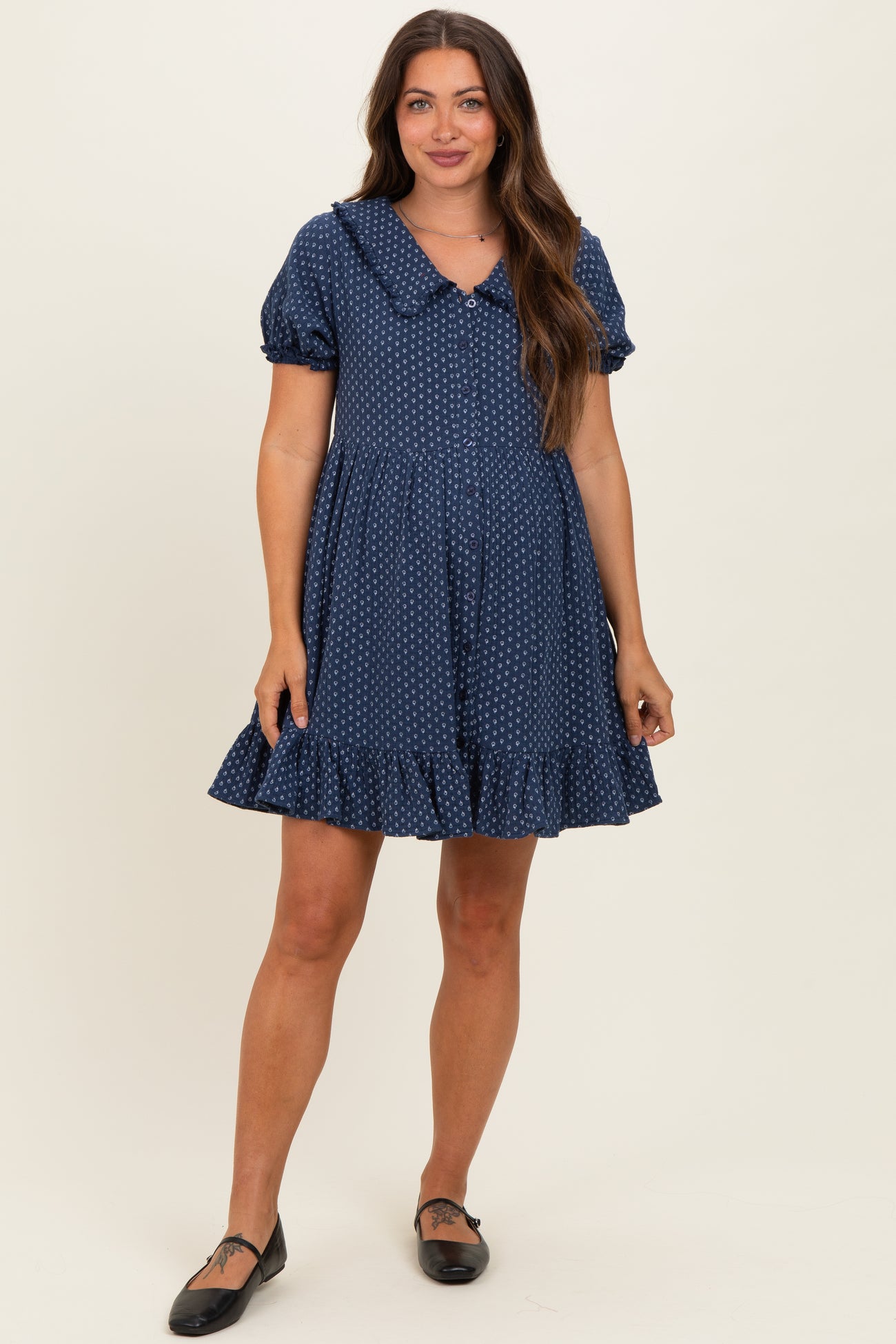 woman wearing a navy blue polka dot maternity dress with a collar and ruffled hem, standing and smiling
