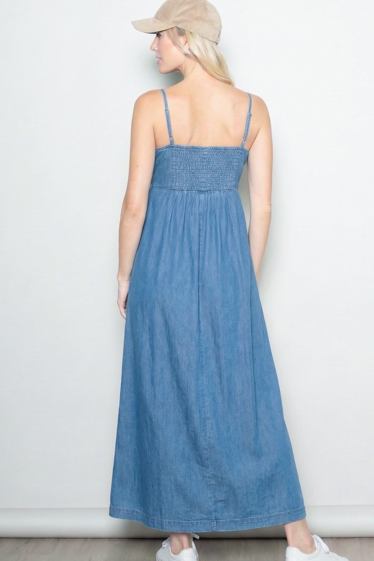 woman wearing a back view of a denim maxi dress with smocked bodice and straps, paired with a beige cap