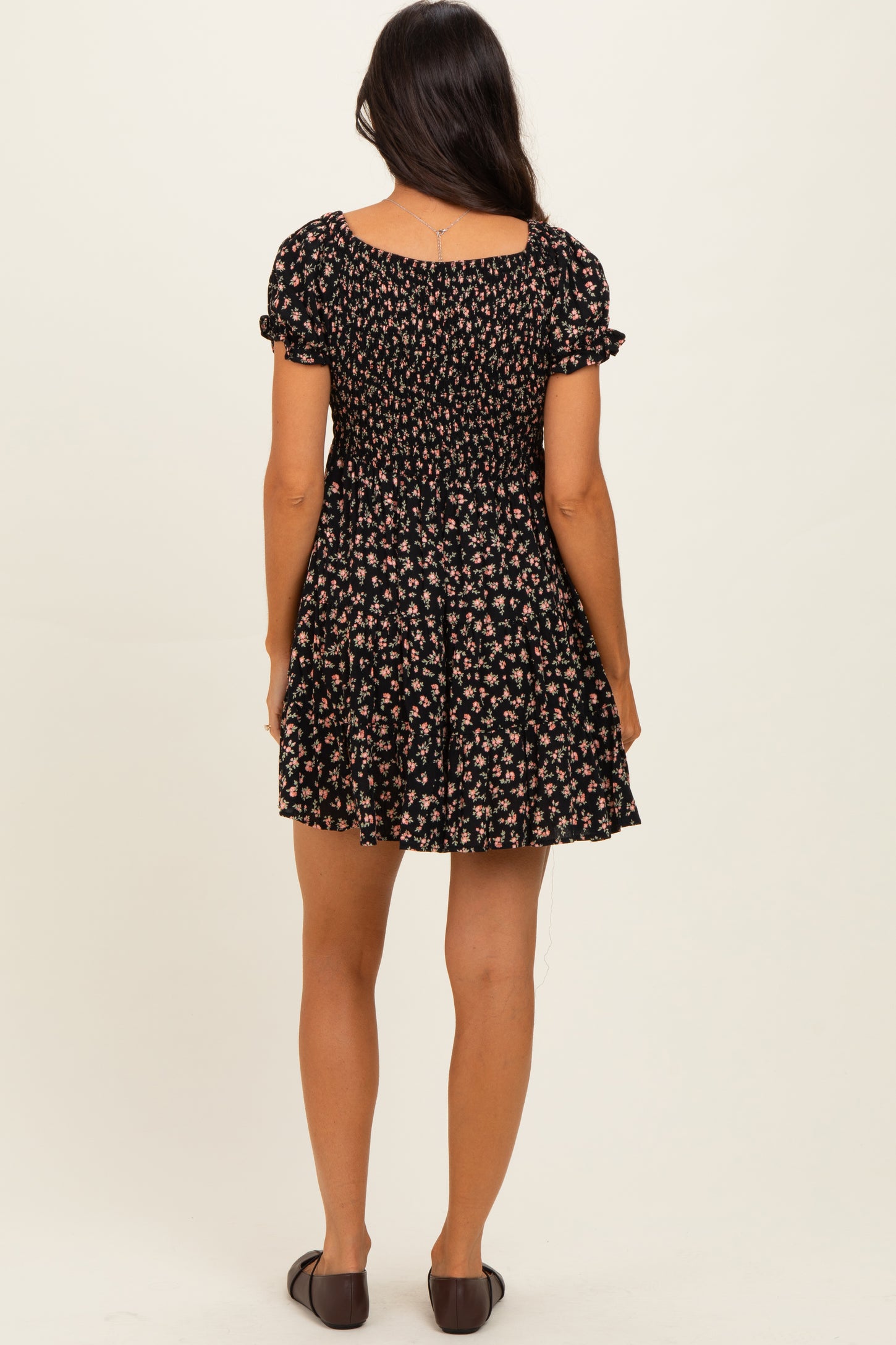 woman wearing a black floral mini dress with short sleeves and a smocked bodice, viewed from the back
