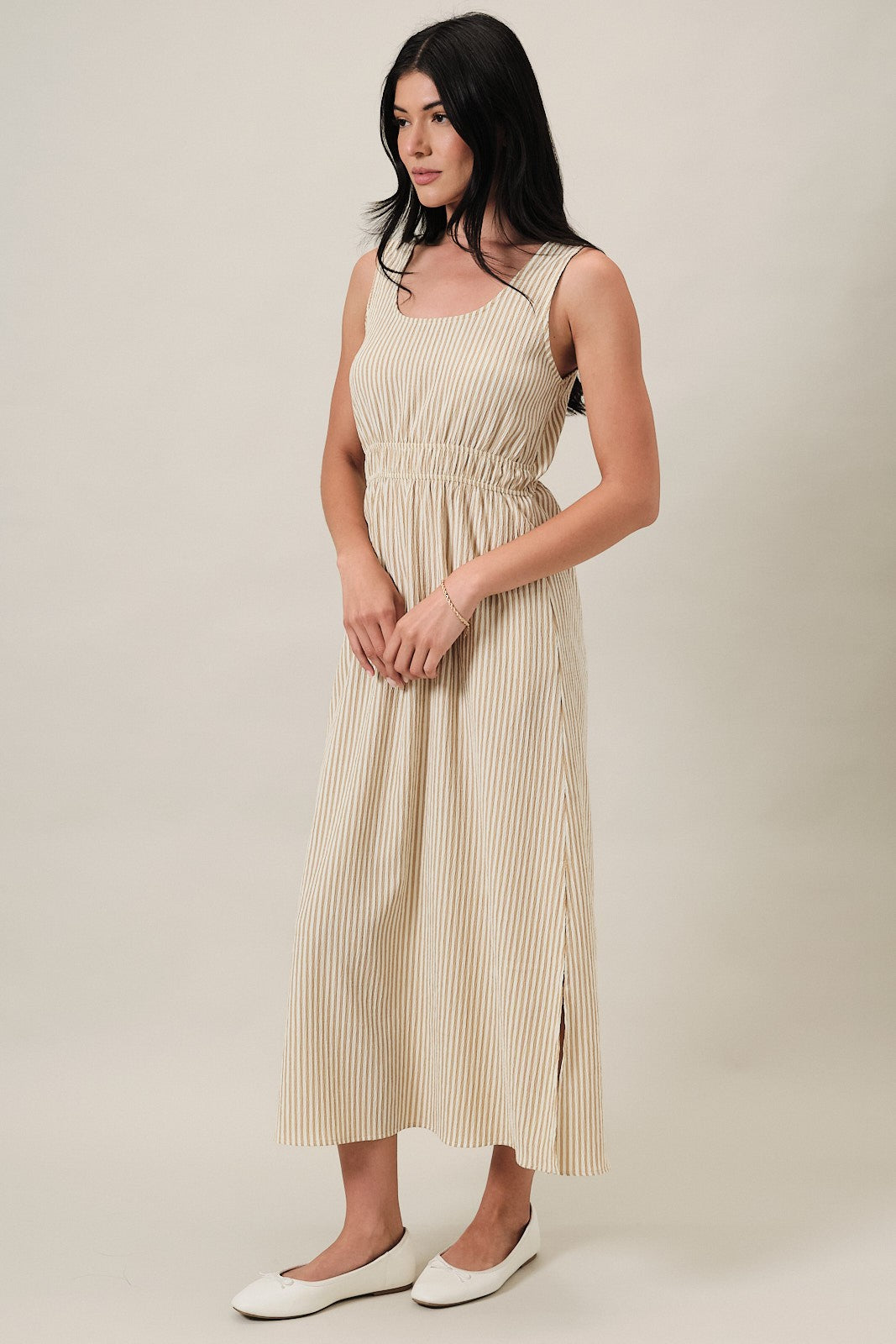 woman wearing a beige striped sleeveless maxi dress with an elastic waistband and side slit, standing on a neutral background