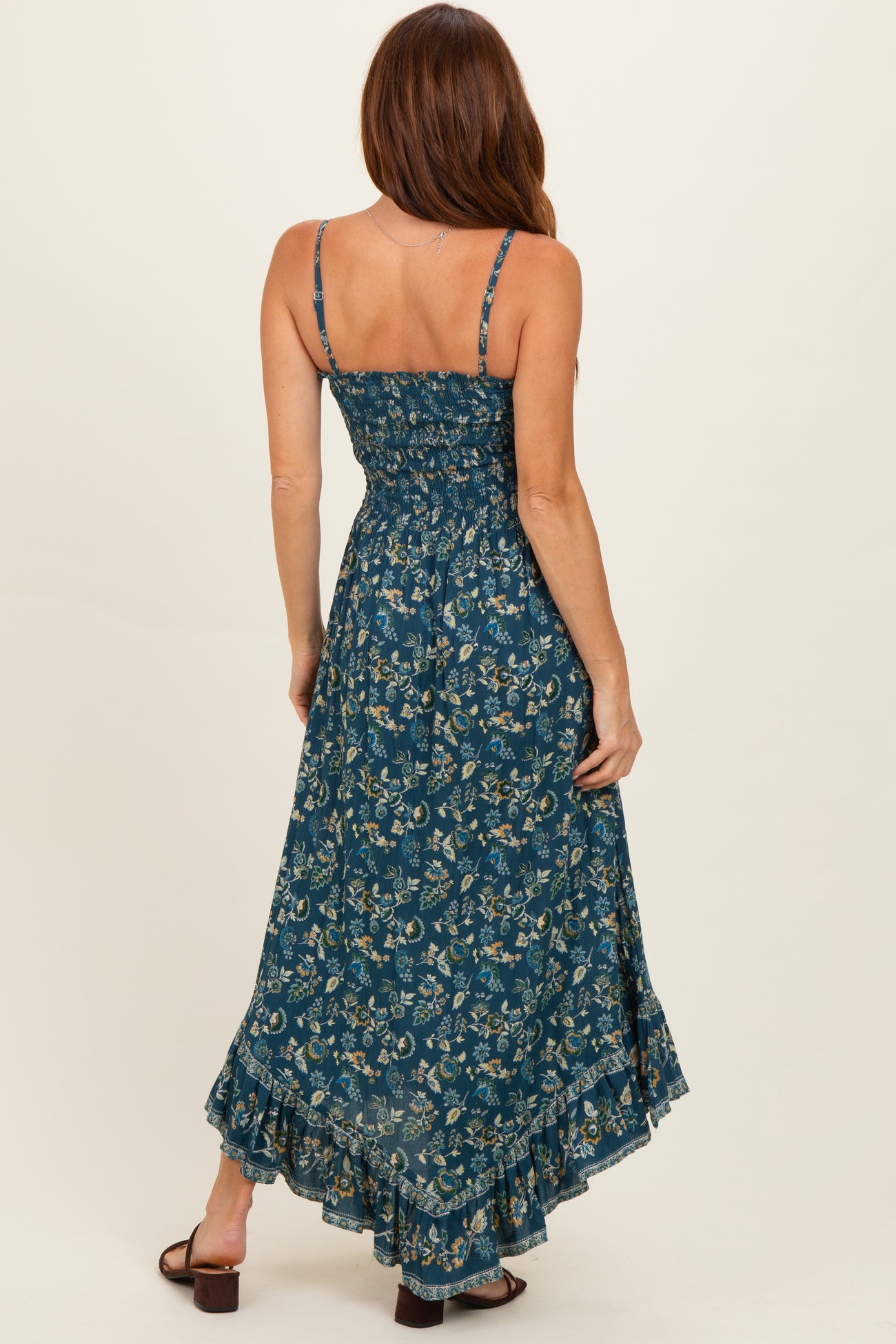 woman wearing a dark teal floral maxi dress with adjustable straps and a ruffled hem, showing the back view