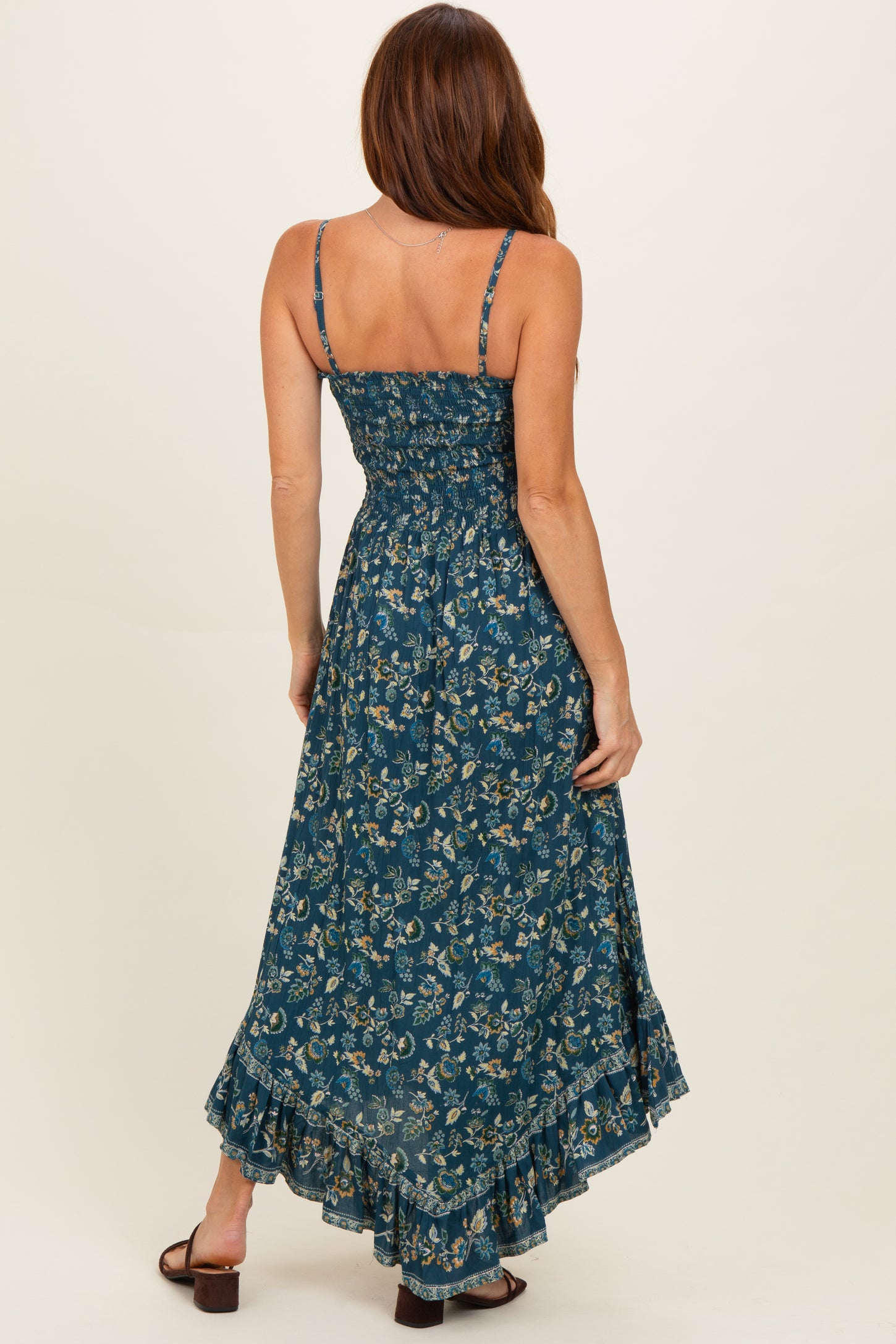 woman wearing a dark teal floral maxi dress with adjustable straps and a ruffled hem, showing the back view