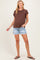 woman wearing a brown maternity top and denim shorts, smiling with hands in pockets, showcasing her baby bump