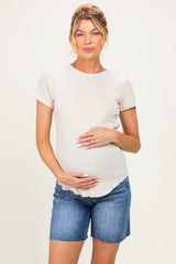 woman wearing a light beige maternity short sleeve top and denim shorts, gently cradling her baby bump