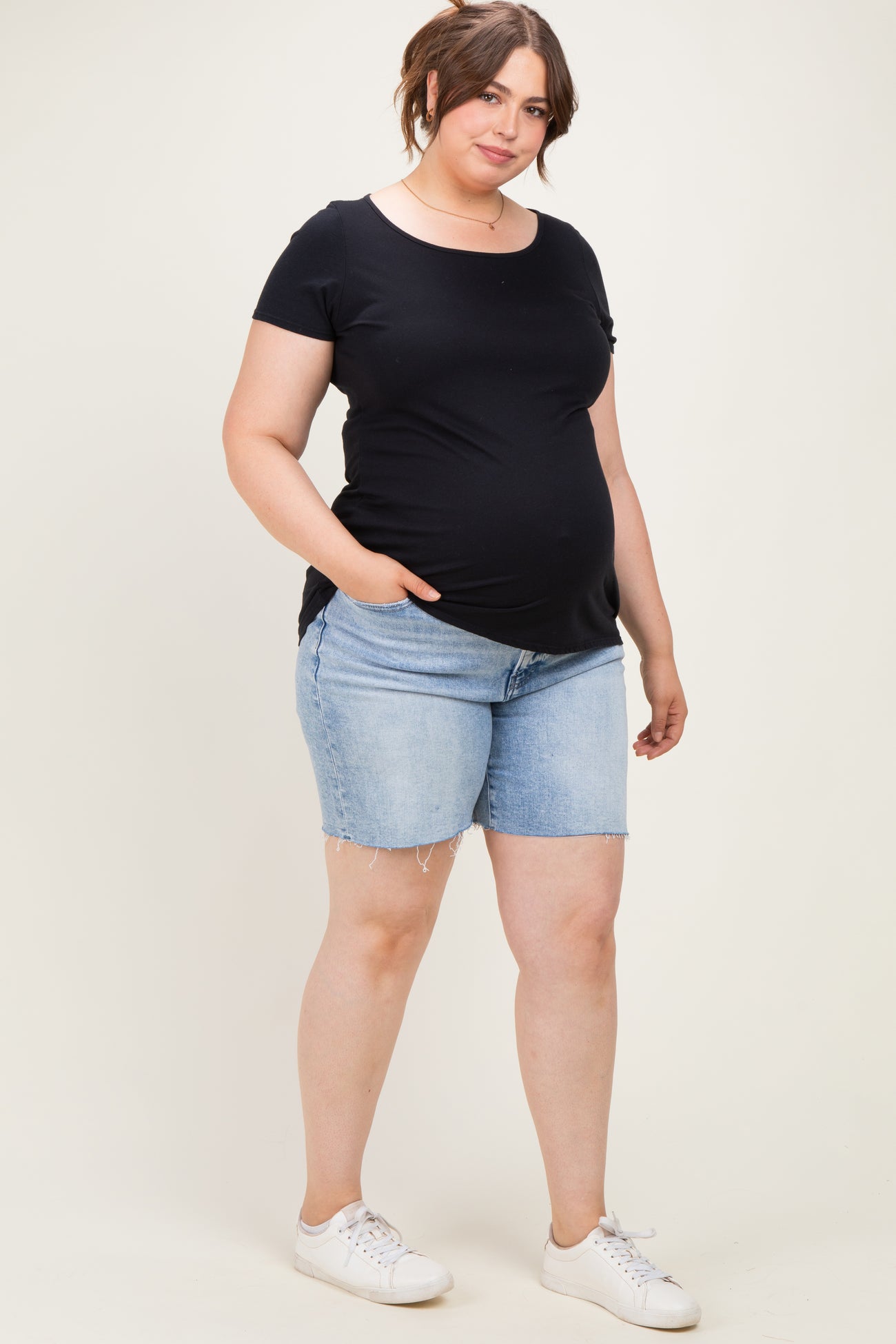 woman wearing a black short sleeve maternity top and blue denim maternity shorts, showing her baby bump