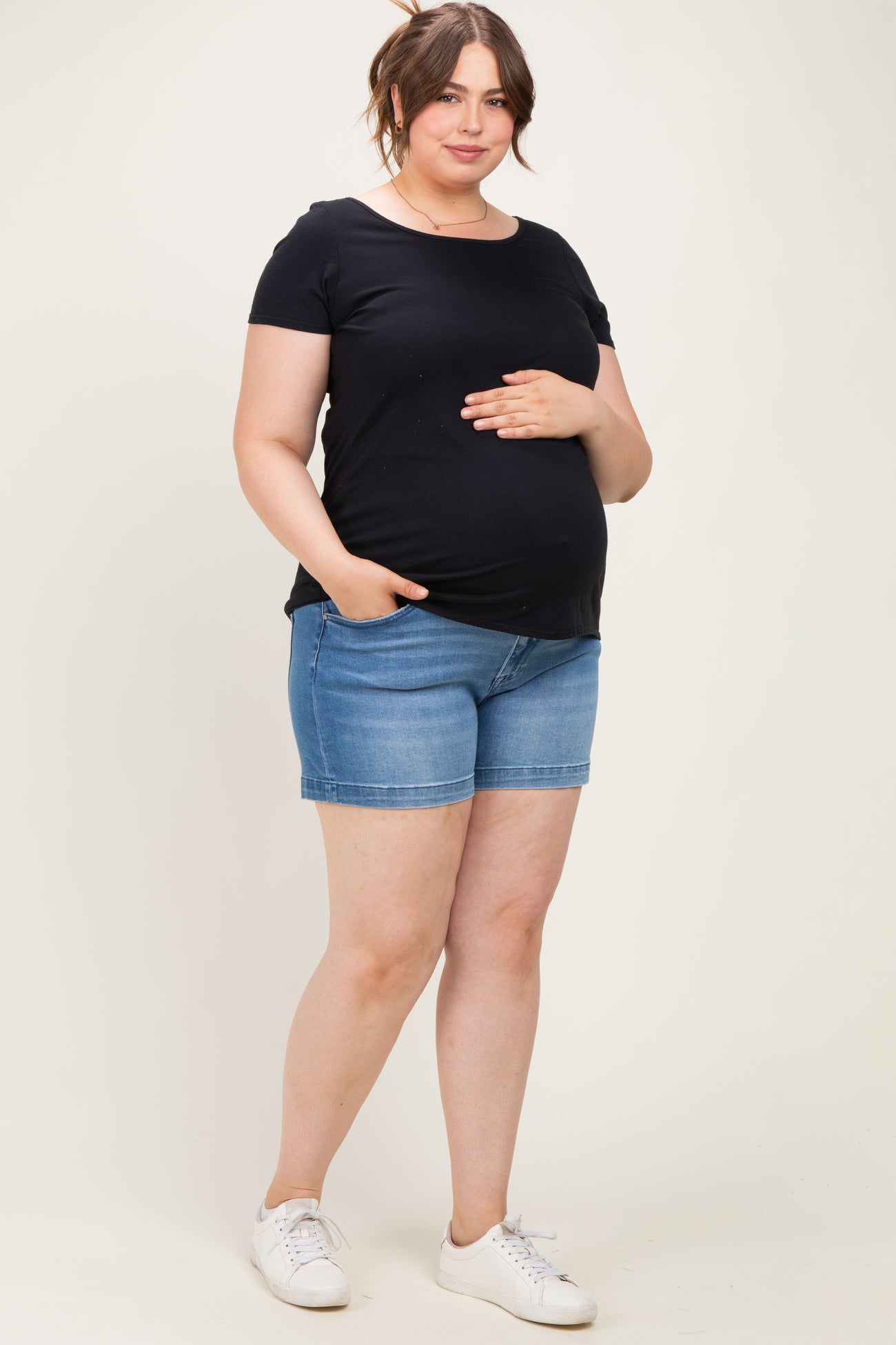 woman wearing a black short sleeve maternity top and denim shorts, holding her baby bump