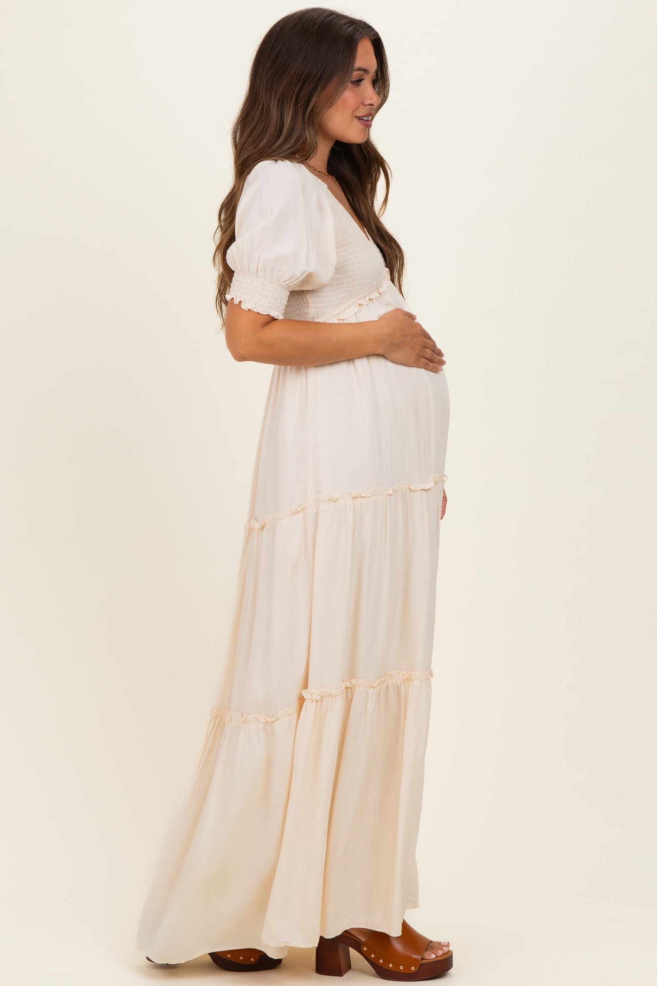 woman wearing a cream maternity maxi dress with puff sleeves and tiered ruffles, gently holding her baby bump