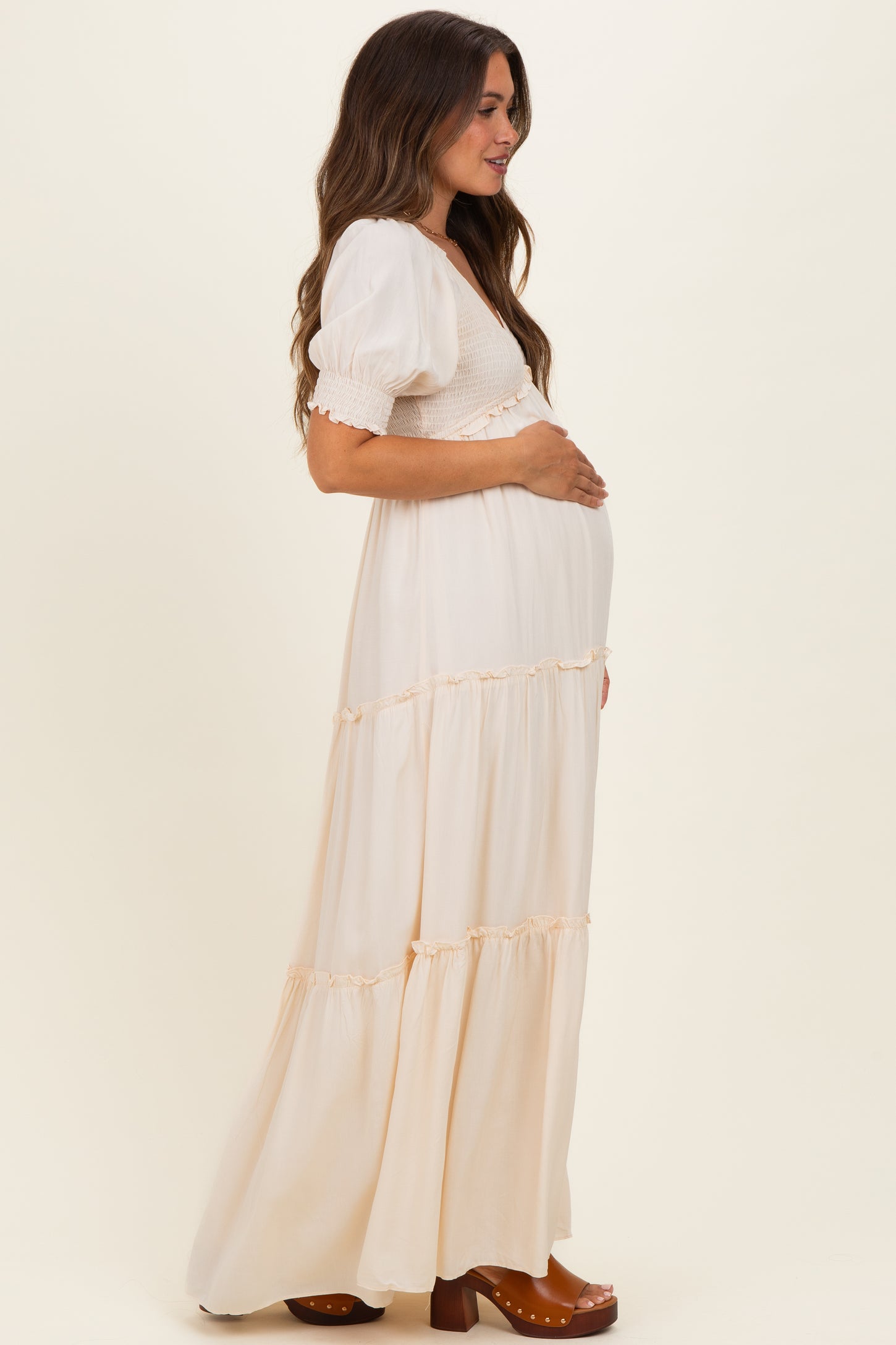 woman wearing a cream maternity maxi dress with puff sleeves and tiered ruffles, gently holding her baby bump