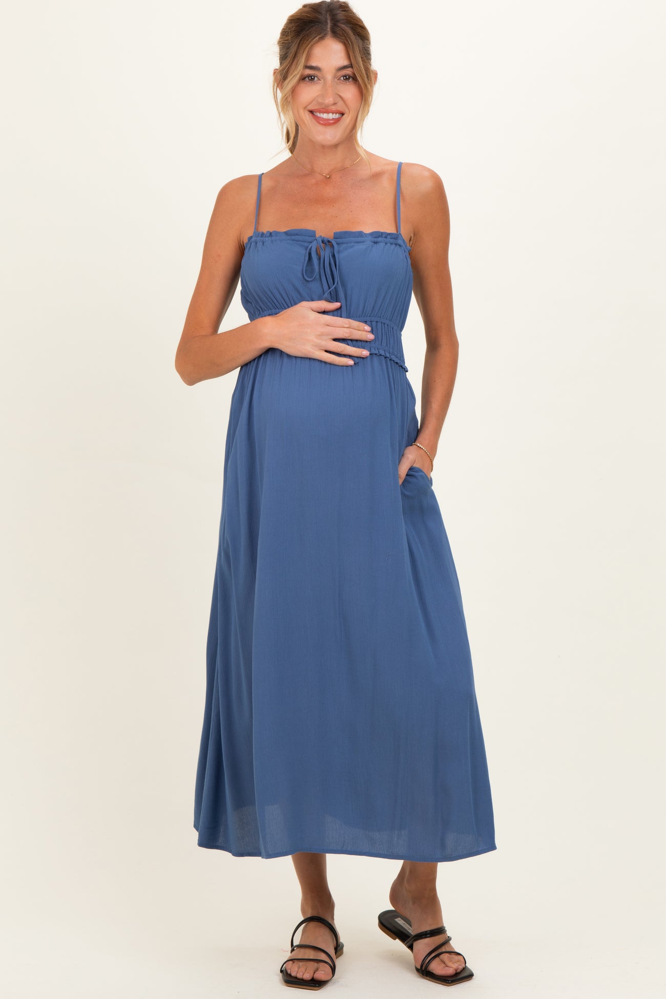 woman wearing a blue sleeveless maternity maxi dress with a tie front and pockets, showcasing her baby bump