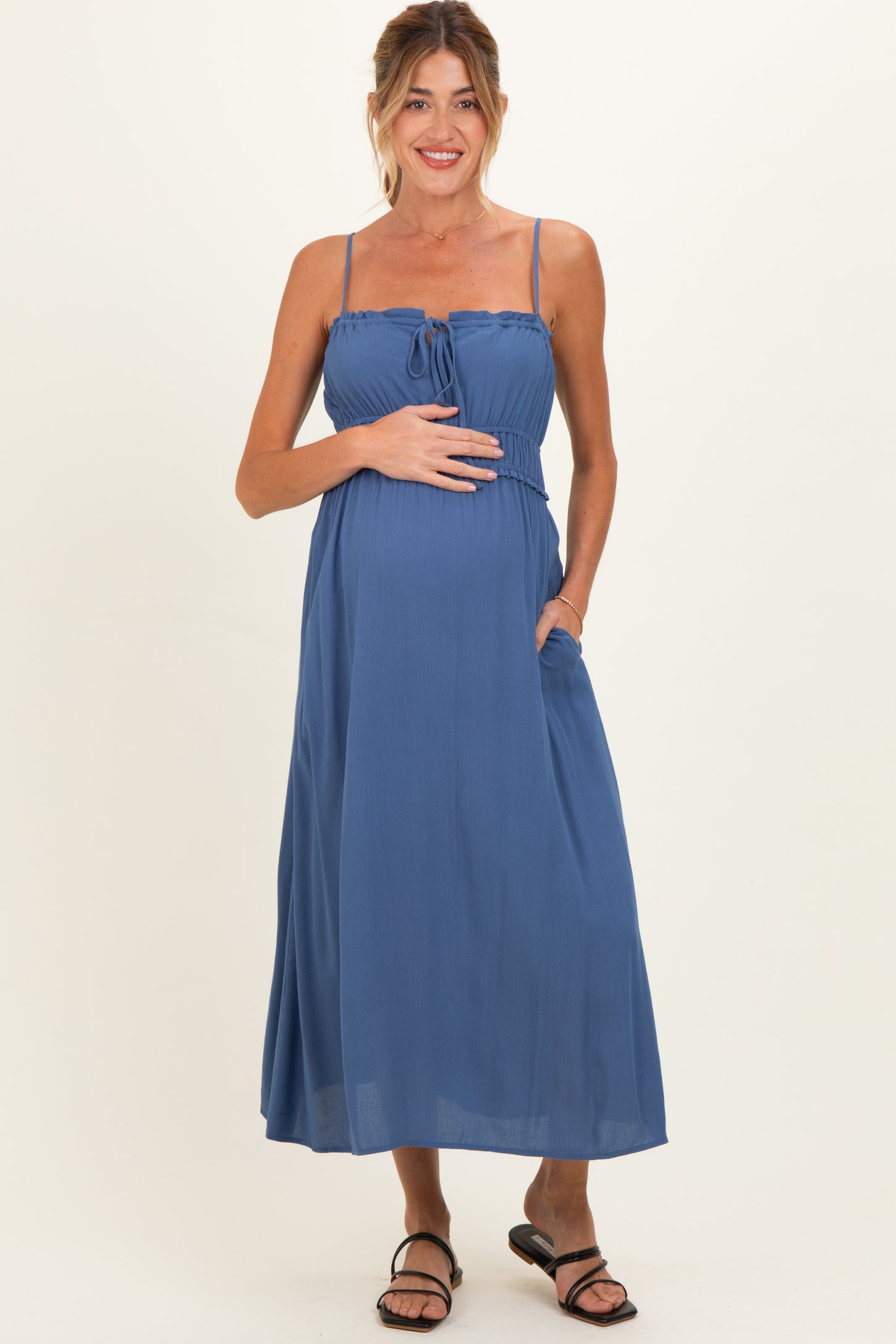 woman wearing a blue sleeveless maternity maxi dress with a tie front and pockets, showcasing her baby bump