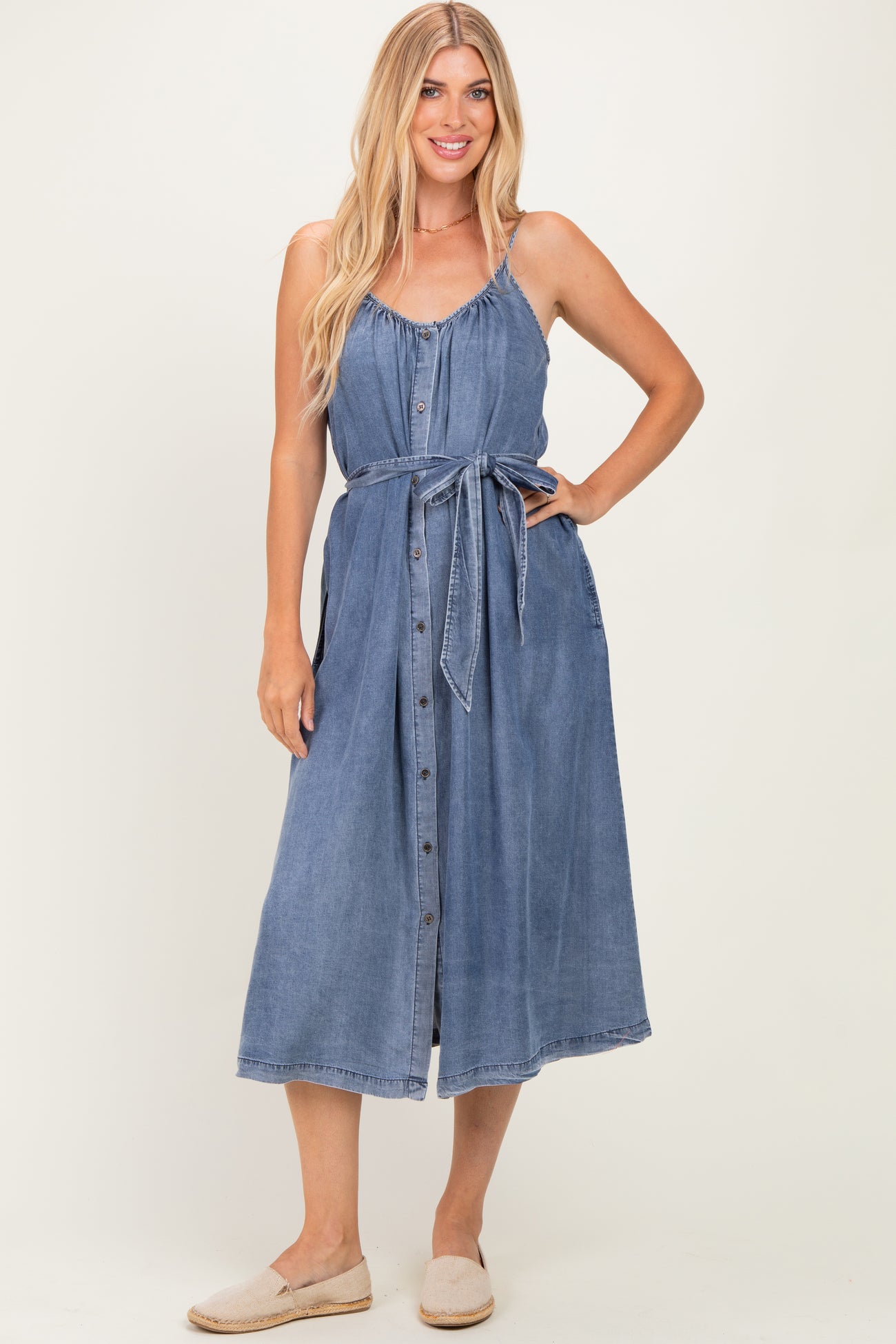 woman wearing a denim midi dress with adjustable straps, a button-down front, and a tied waist belt