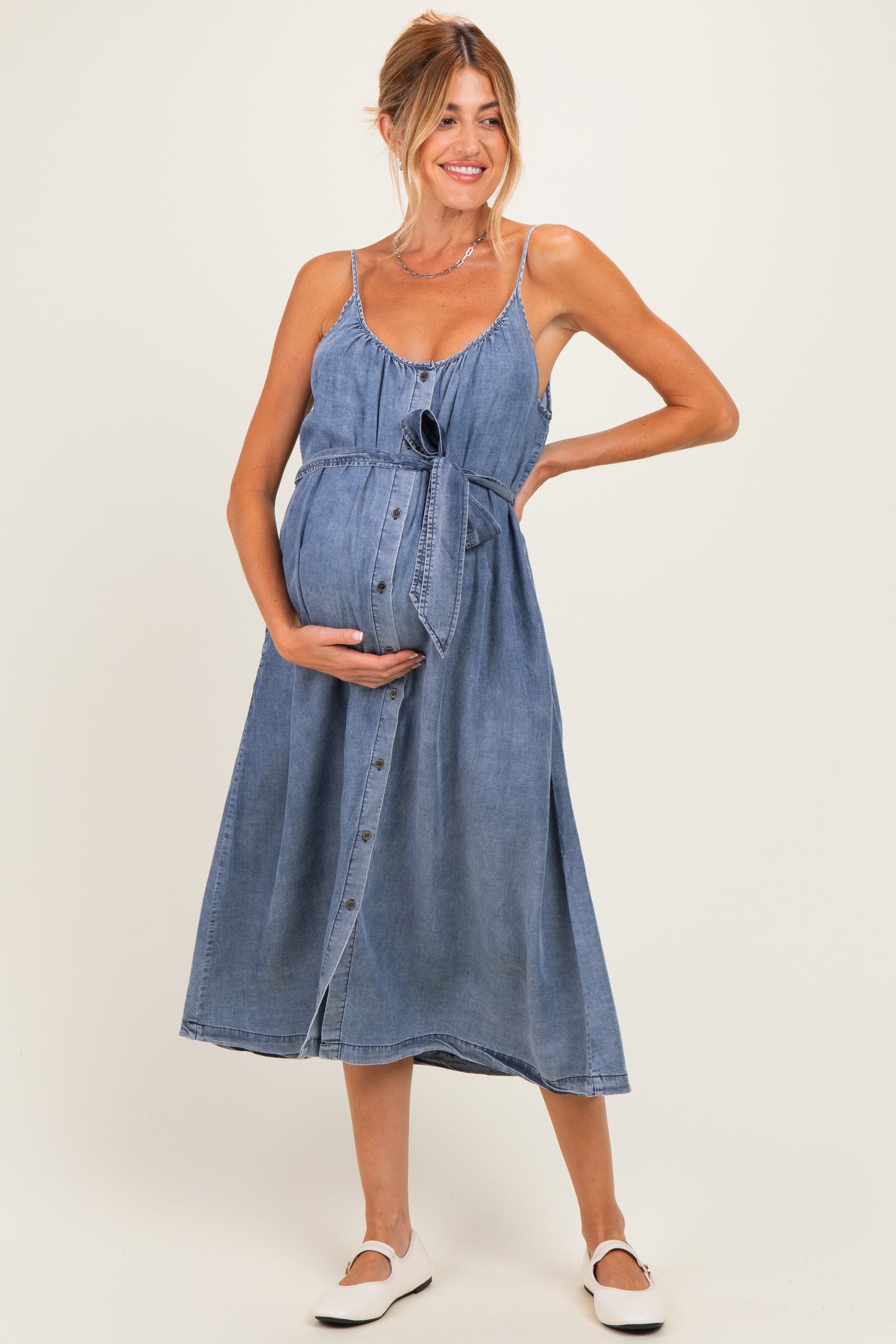 woman wearing a blue denim maternity dress with adjustable straps and a tie at the waist, showcasing her baby bump