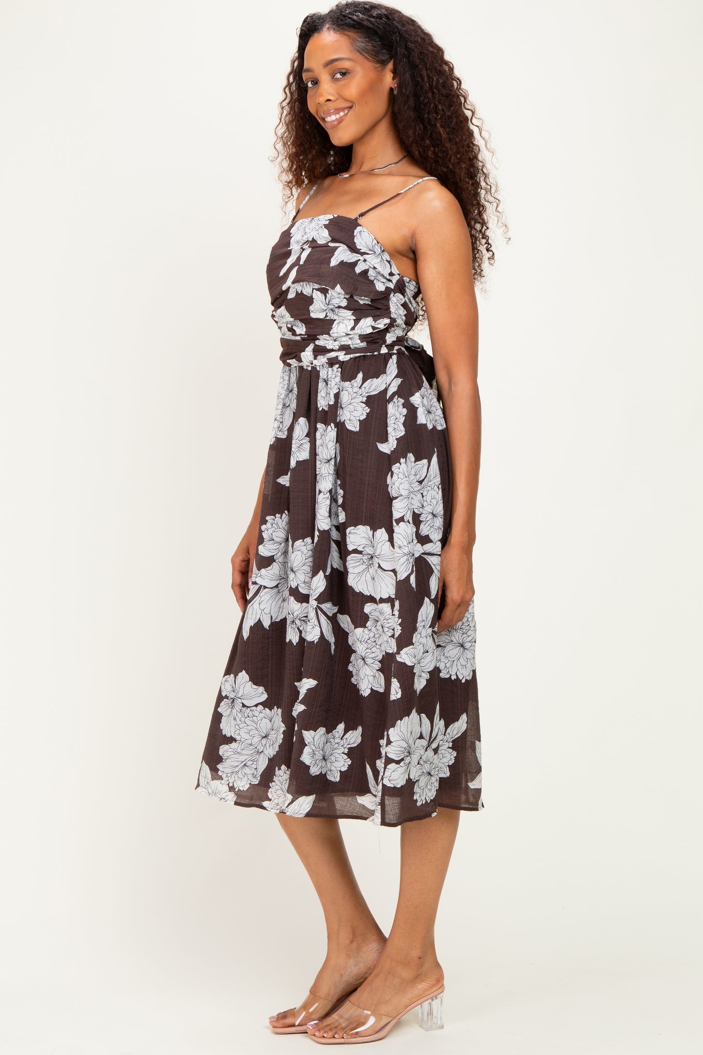 woman wearing a dark brown floral midi dress with adjustable straps and a fitted waist, standing against a light background