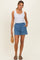woman wearing a white sleeveless henley top and blue denim shorts, holding a woven bag