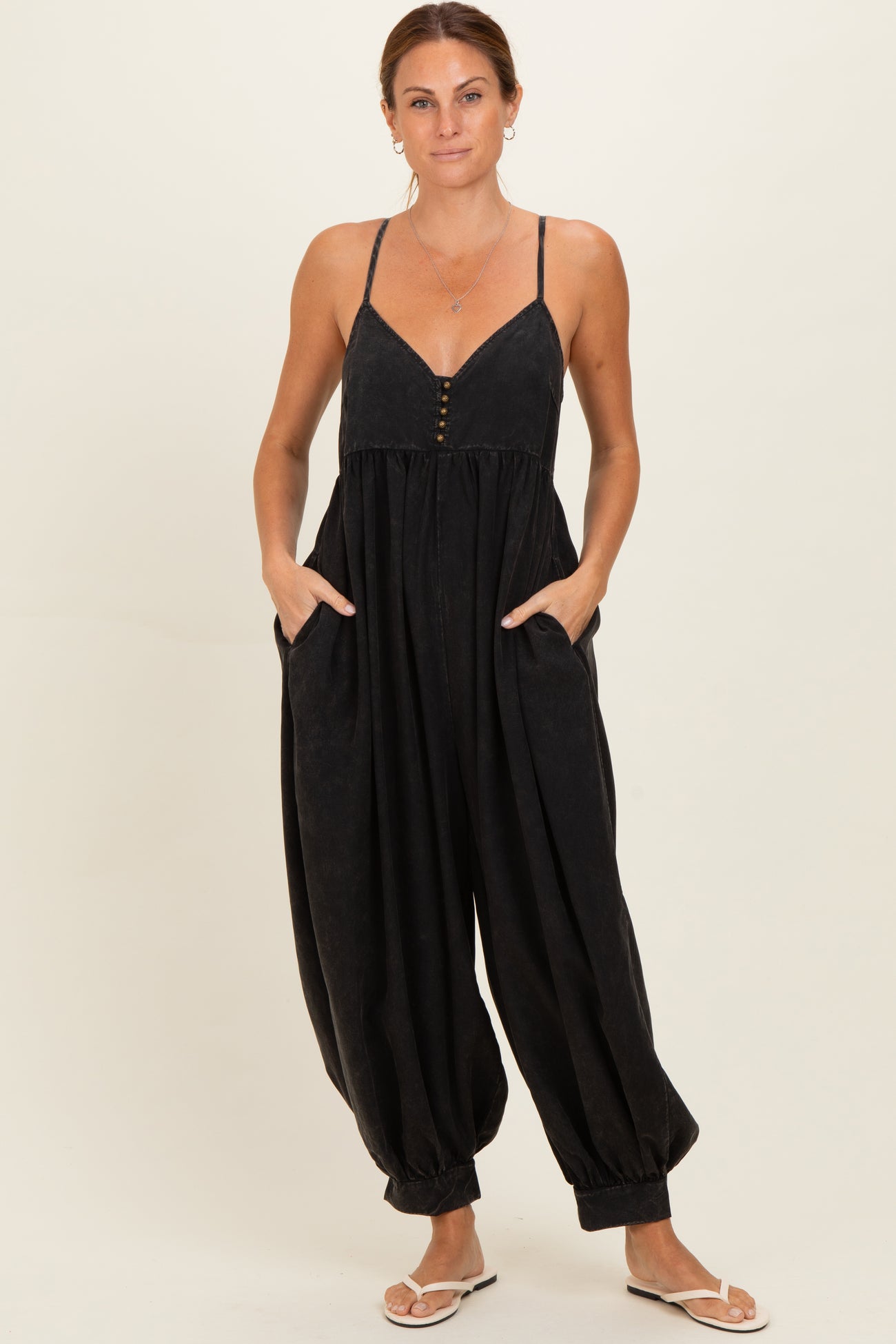 woman wearing a black spaghetti strap jumpsuit with button details and pockets, styled with flat sandals
