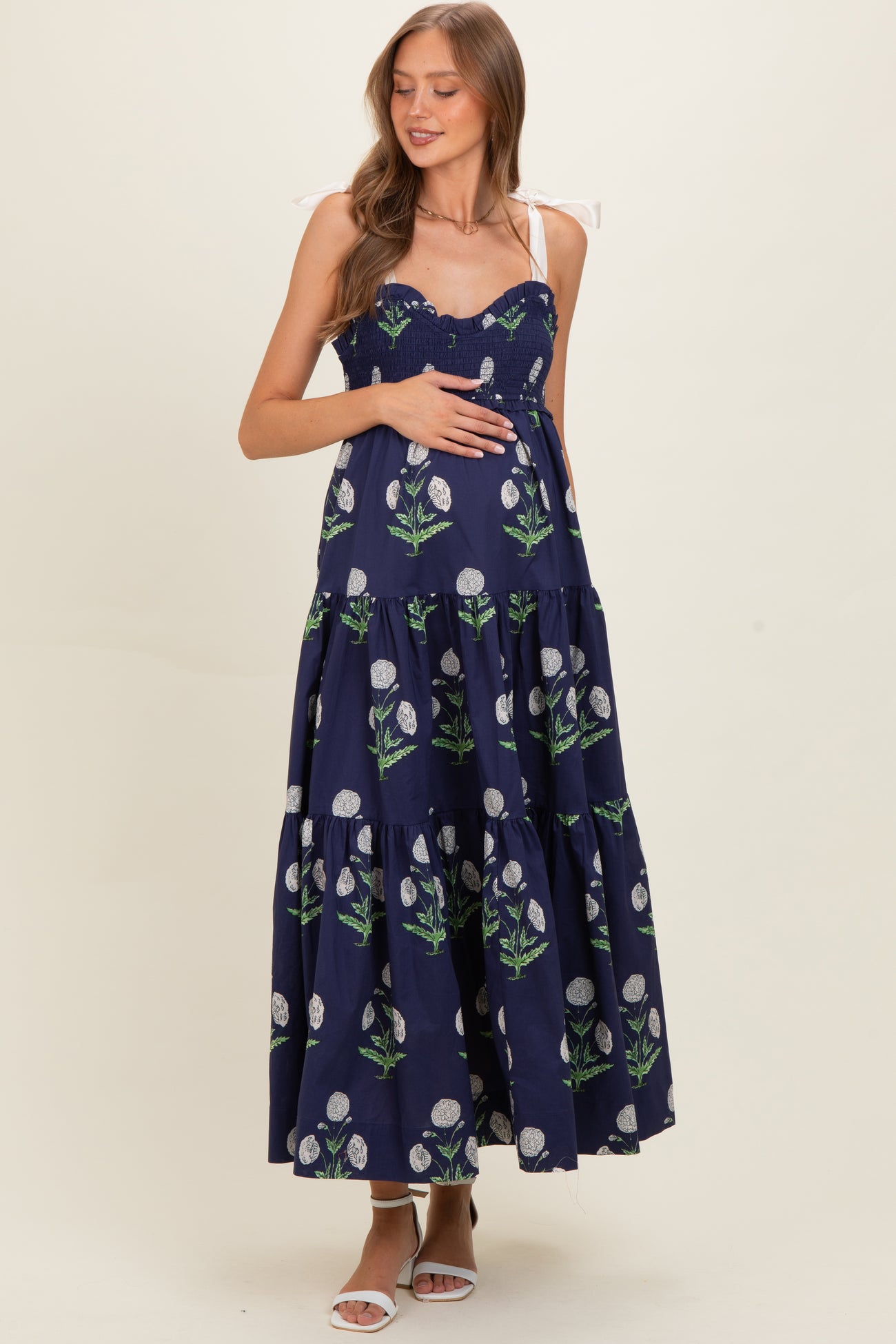 woman wearing a navy blue floral print maternity maxi dress with smocked bodice and adjustable straps, holding her baby bump