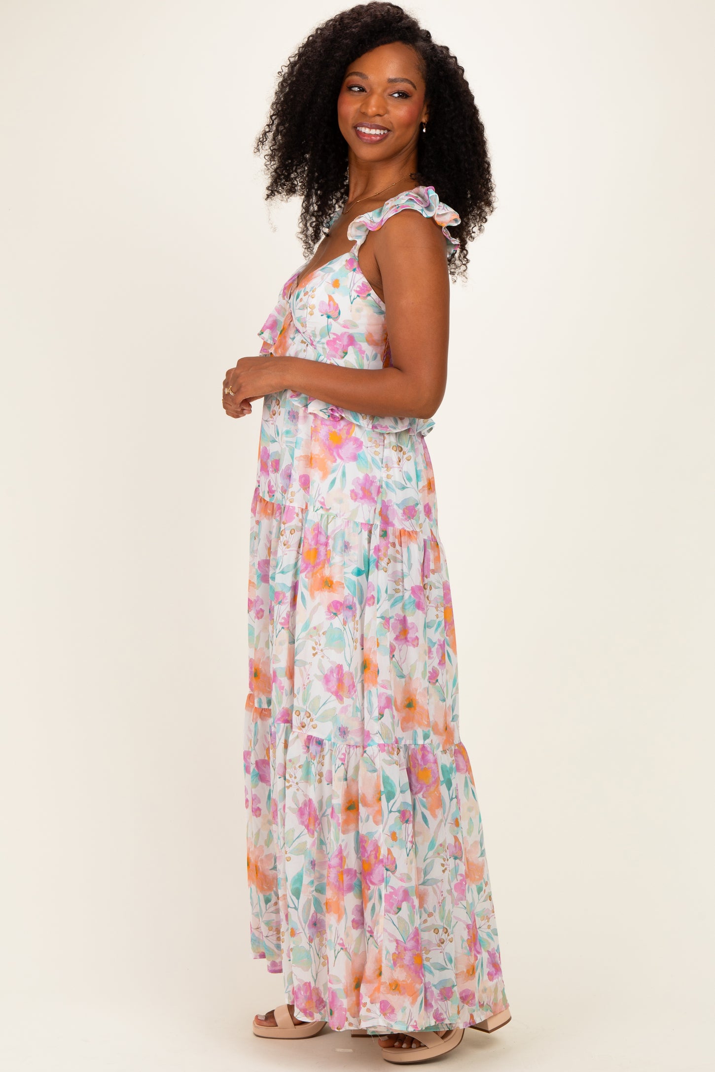 woman wearing a flowy maxi dress with colorful floral prints and ruffled straps, standing sideways against a light background