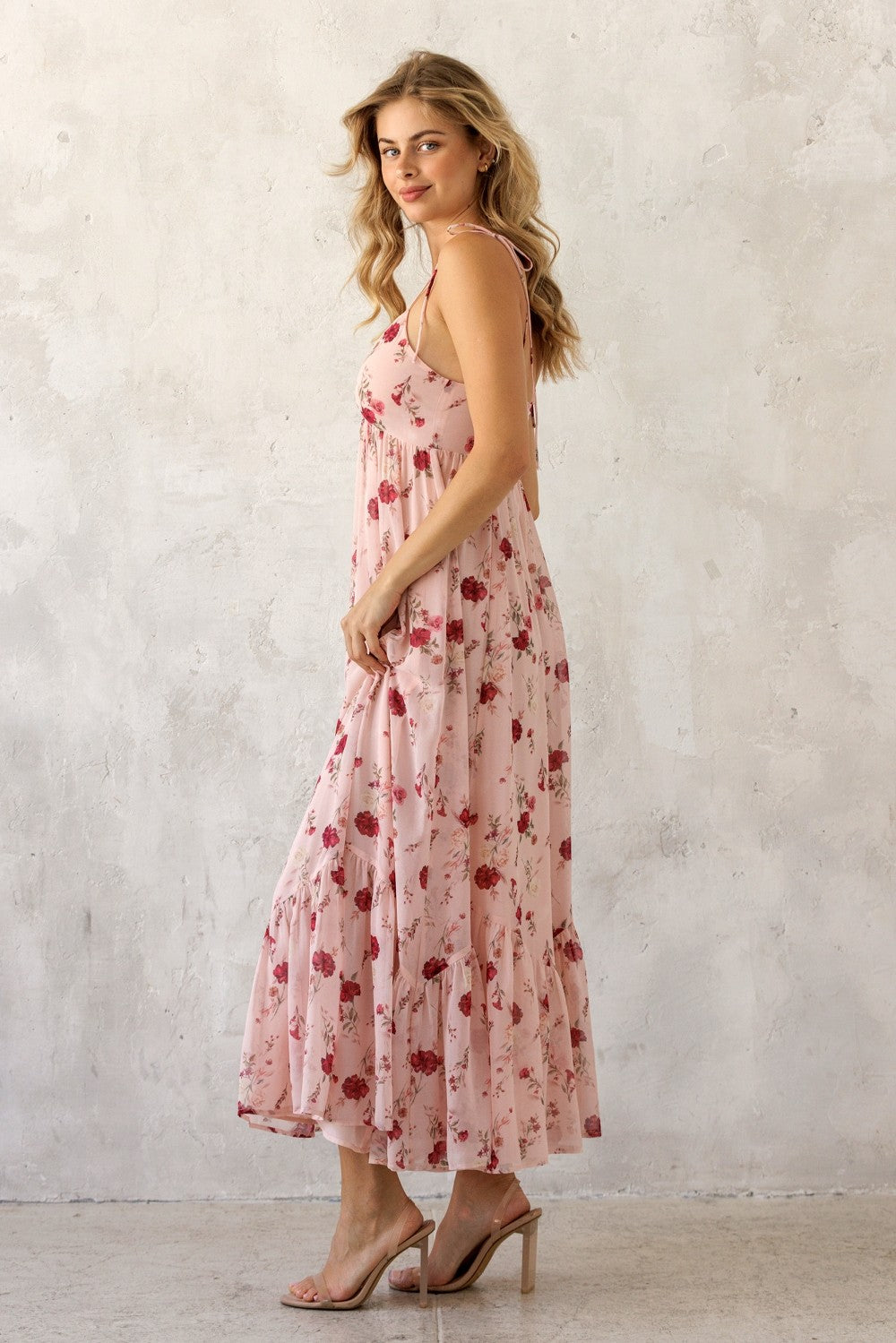 woman wearing a pink floral maxi dress with adjustable straps and a tiered skirt, posing sideways against a textured wall