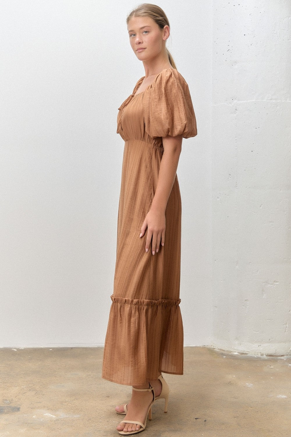 woman wearing a brown puff sleeve maxi dress with a fitted waist and ruffled hem, standing in a side profile