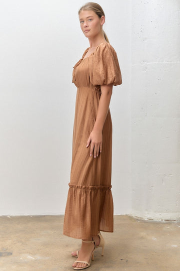 woman wearing a brown puff sleeve maxi dress with a fitted waist and ruffled hem, standing in a side profile