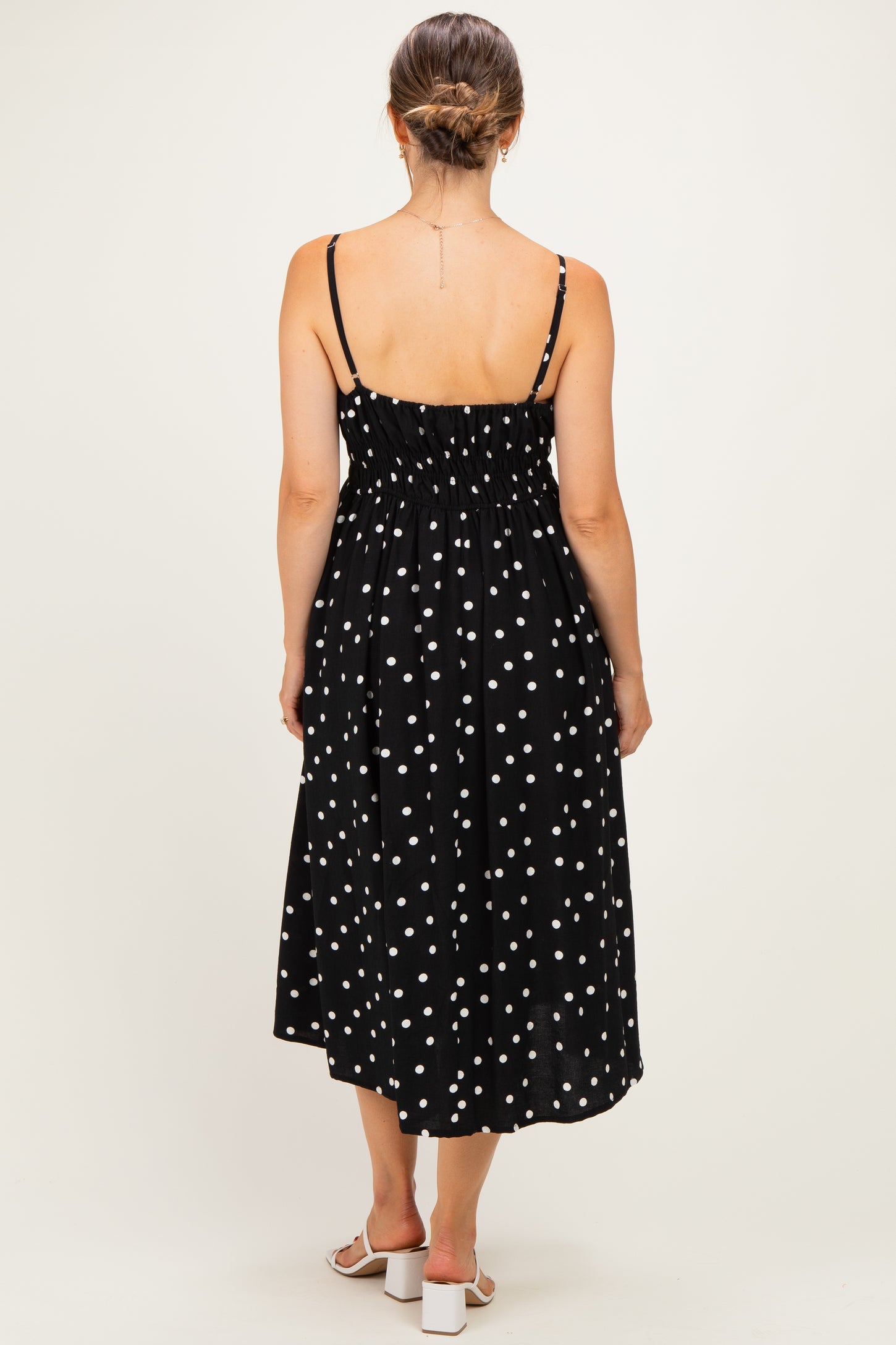 woman wearing a black polka dot maternity midi dress with adjustable straps and a smocked back, view from the back