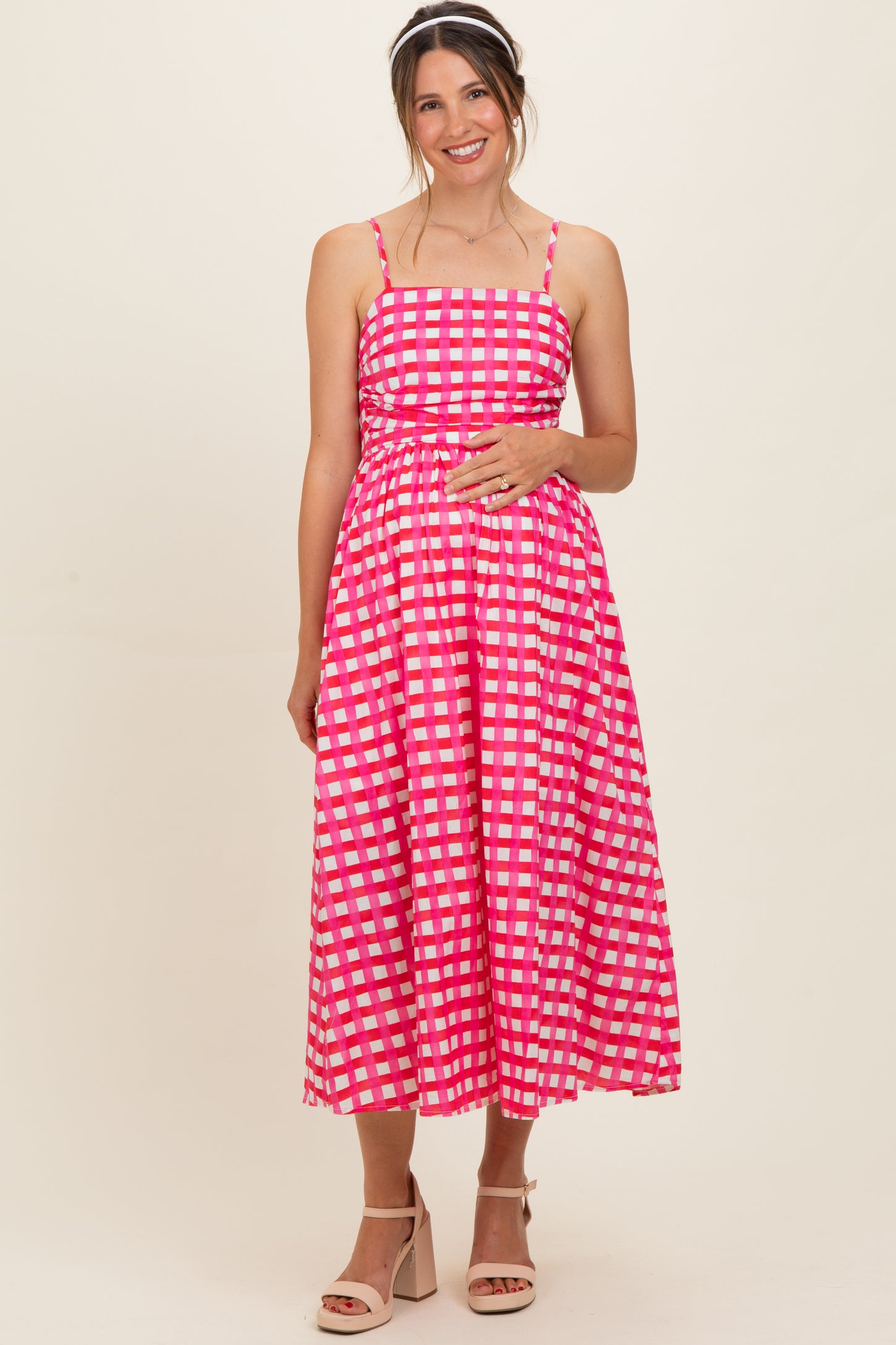 woman wearing a pink and white checked maternity dress with adjustable spaghetti straps and a fitted bodice, smiling with hands on her baby bump