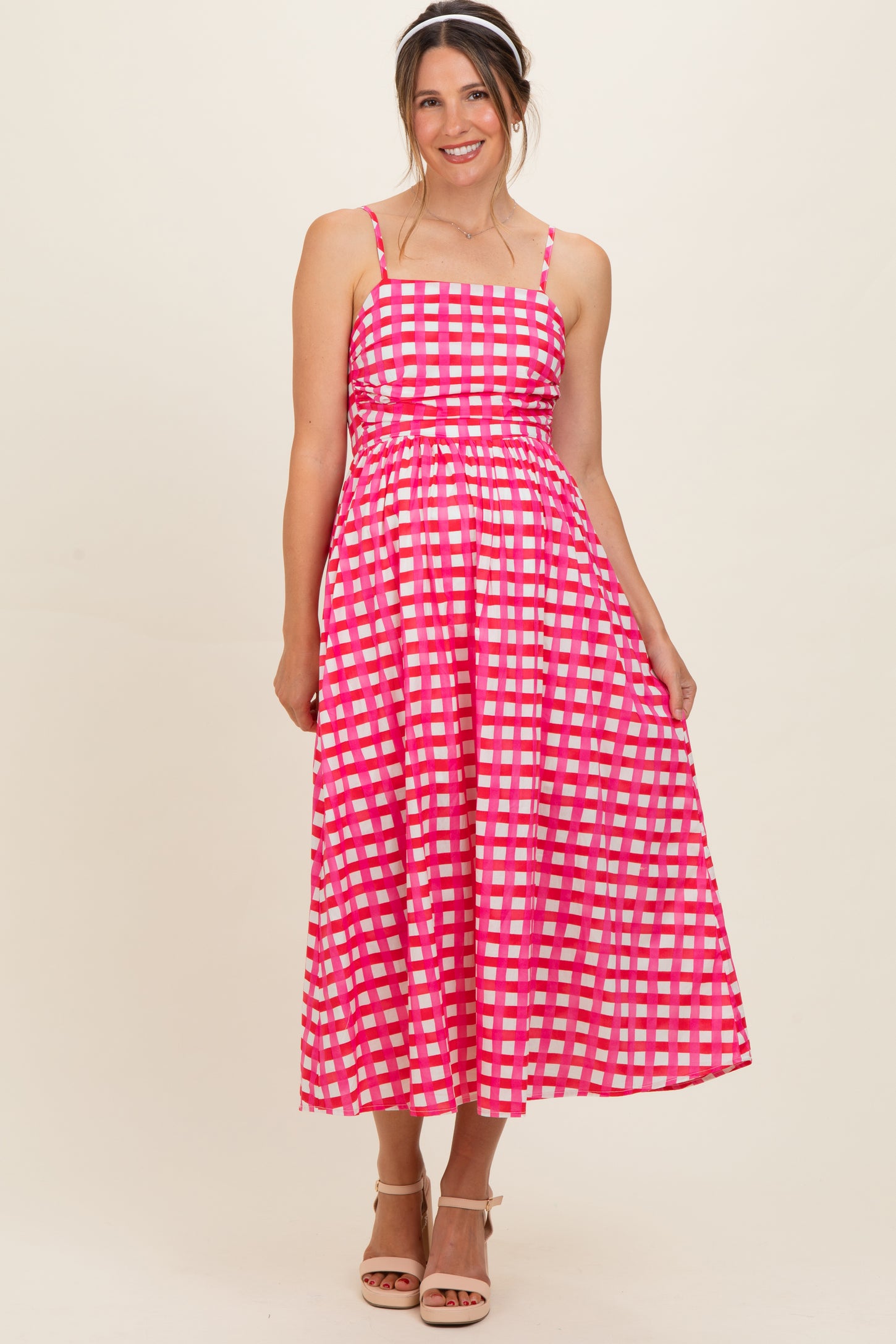woman wearing a pink checkered maternity maxi dress with spaghetti straps and a fitted bodice, smiling at the camera