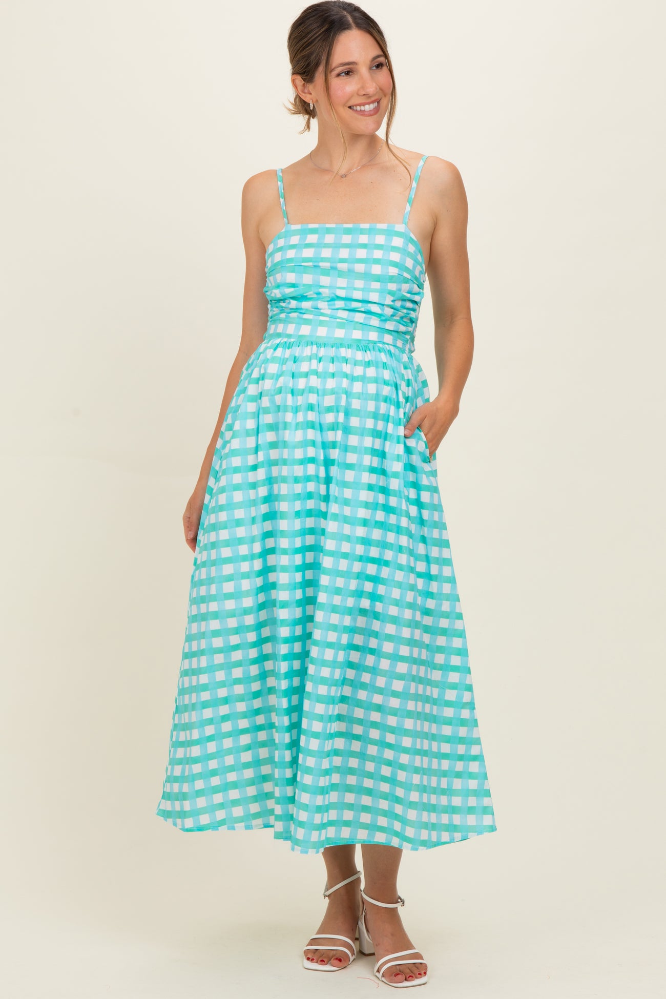 woman wearing a turquoise maternity dress with a checkered pattern and pockets, styled with spaghetti straps