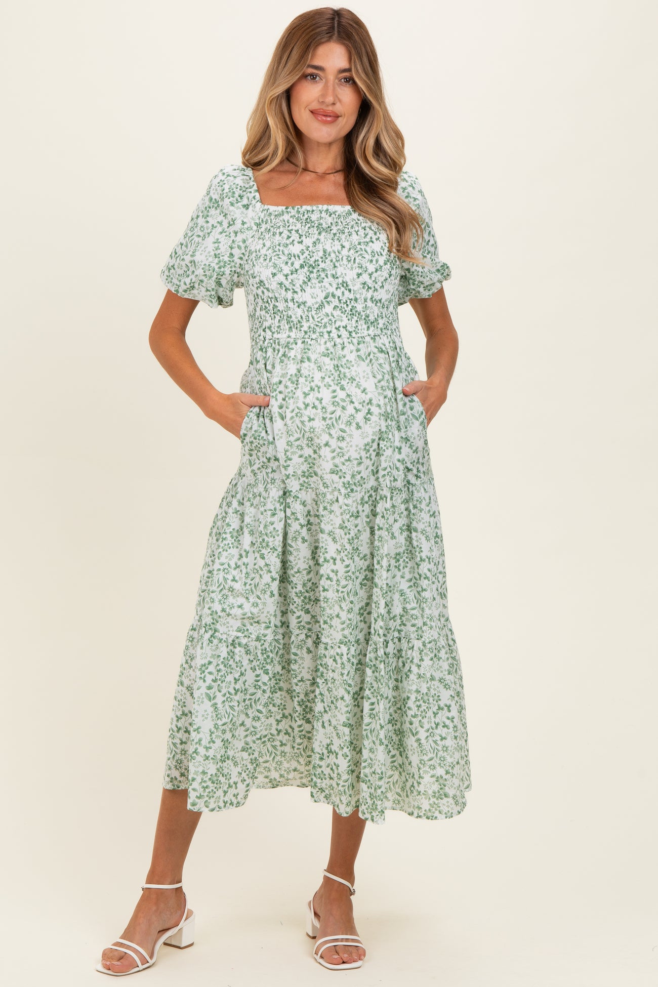 woman wearing a green floral midi maternity dress with puff sleeves and pockets, standing against a neutral background
