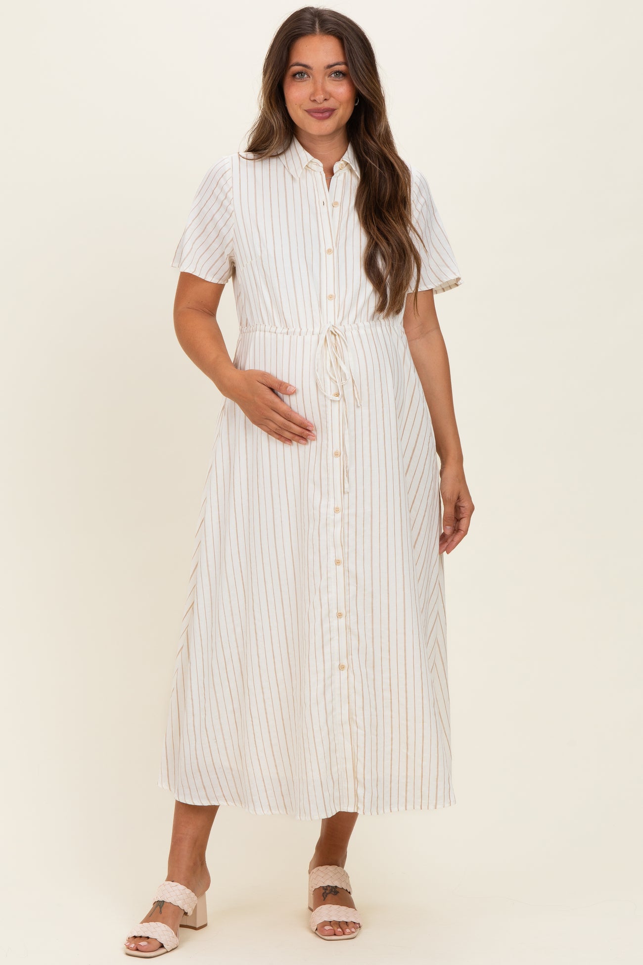 woman wearing a white striped maternity dress with short sleeves and a tie waist, holding her baby bump
