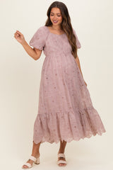 woman wearing a light mauve maternity dress with eyelet detailing and a tiered hem, smiling and showing off her baby bump